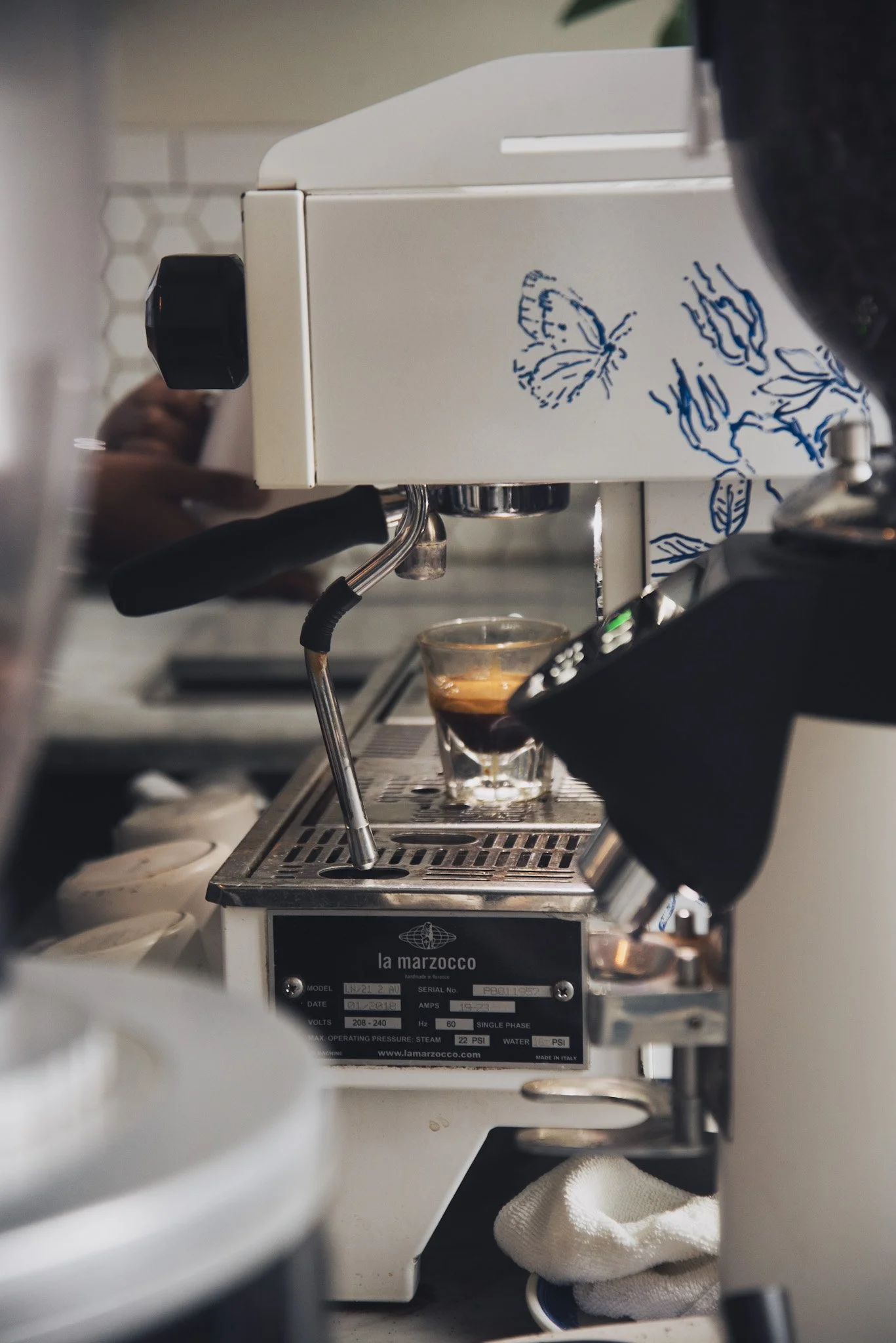Close-up of a white espresso machine with blue butterfly and leaf design, brewing coffee into a glass cup, with parts of the machine and a hand with a white towel visible.
