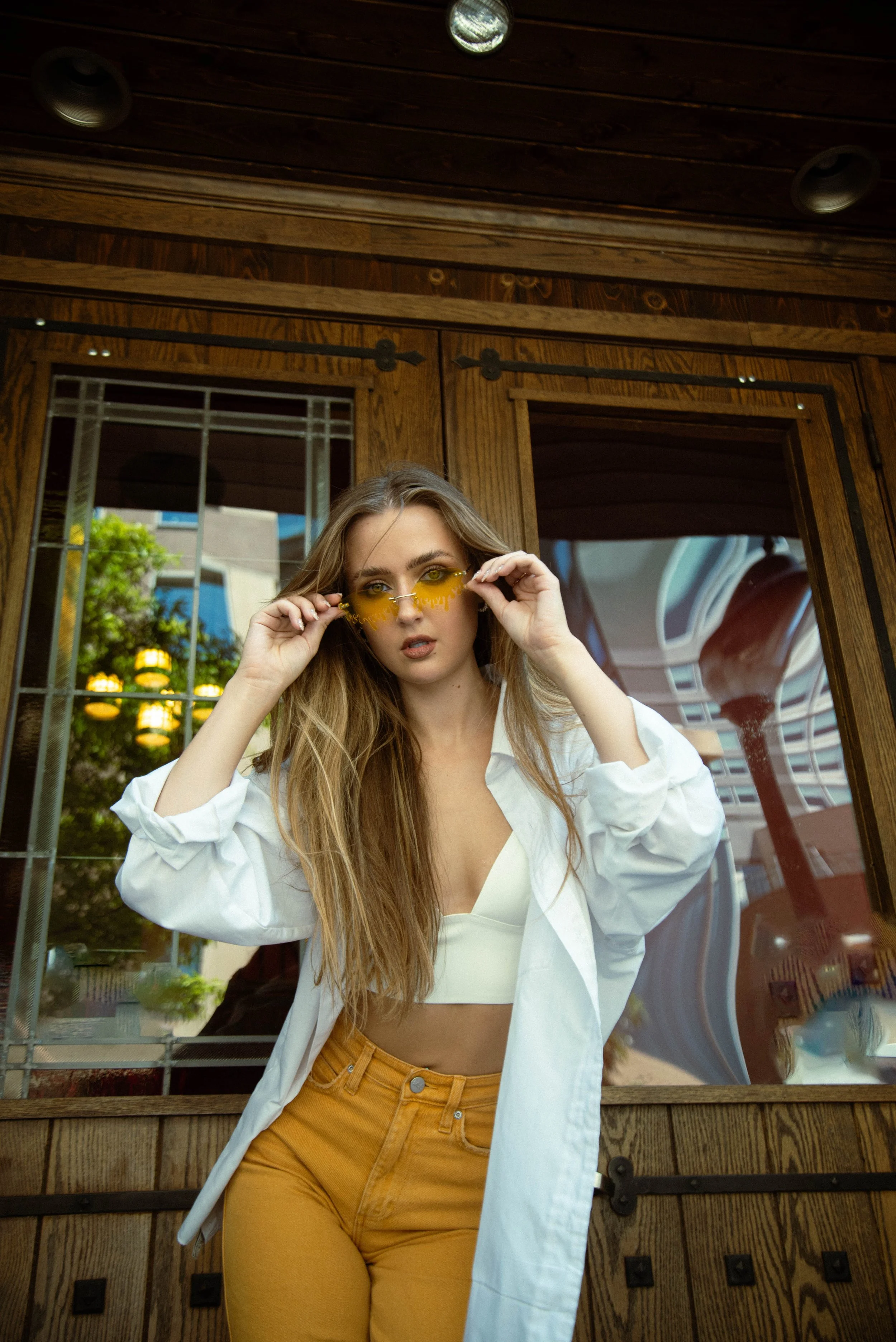A young woman with long blonde hair wearing yellow sunglasses, a white crop top, mustard-colored pants, and an open white shirt, standing in front of a wooden structure and adjusting her sunglasses.