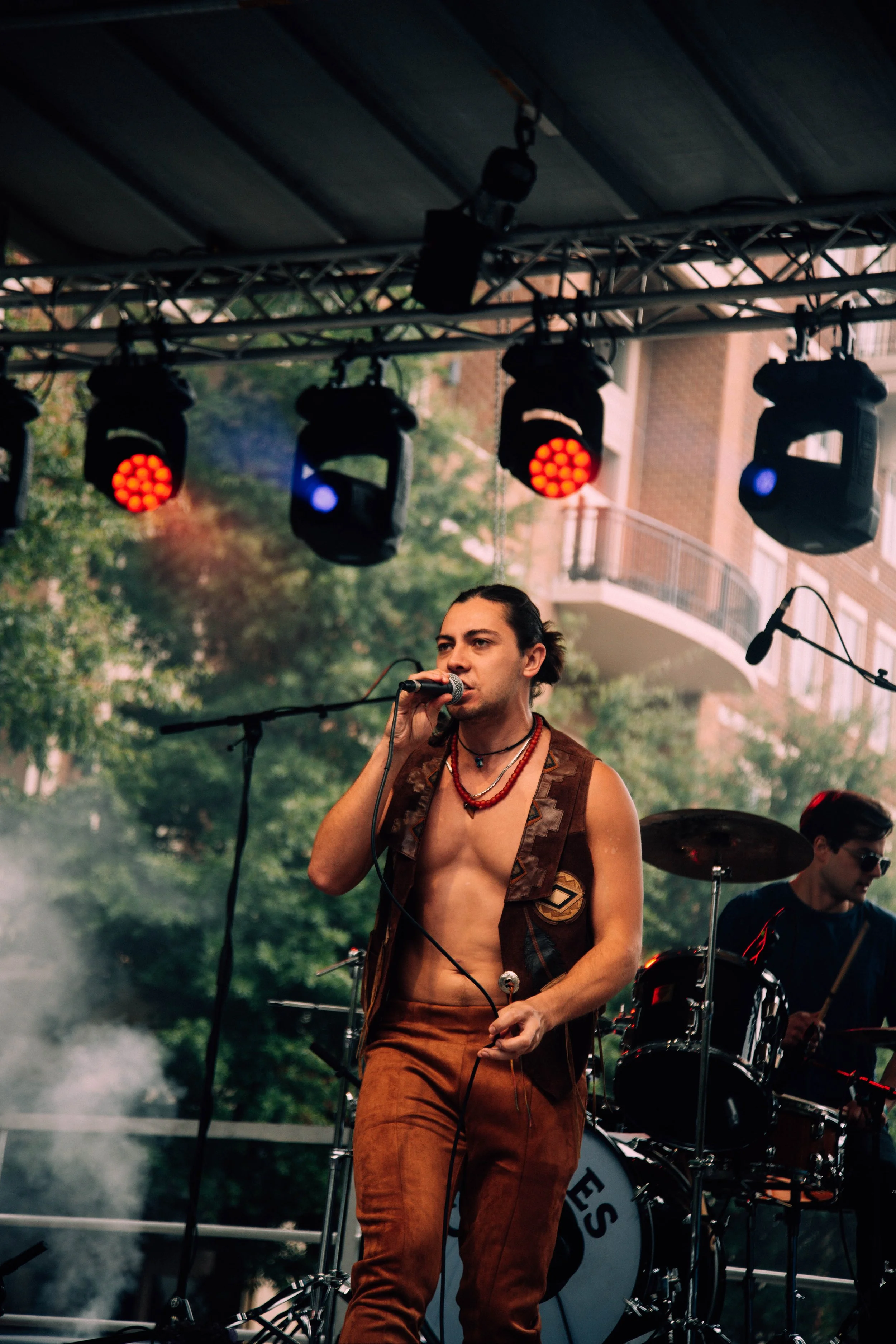 A shirtless man with long hair singing into a microphone during a live outdoor music performance, with drums and other band members in the background, under stage lighting.