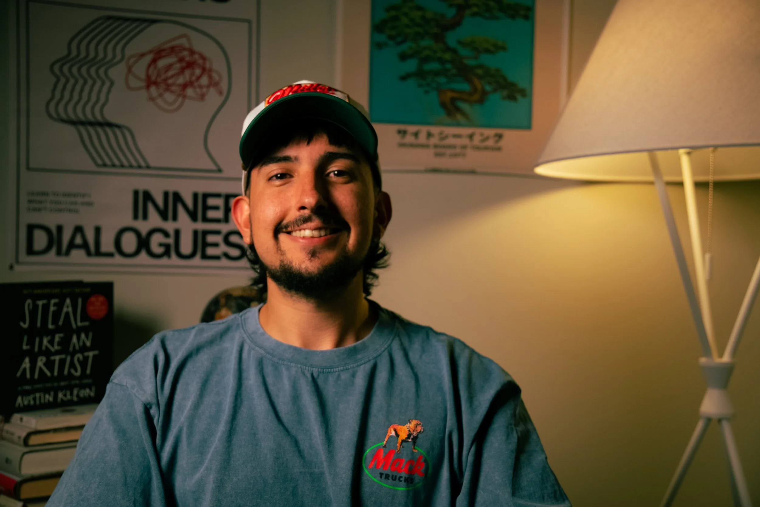 A young man wearing a Mack Trucks T-shirt and a baseball cap smiling in a room with posters and a lamp.
