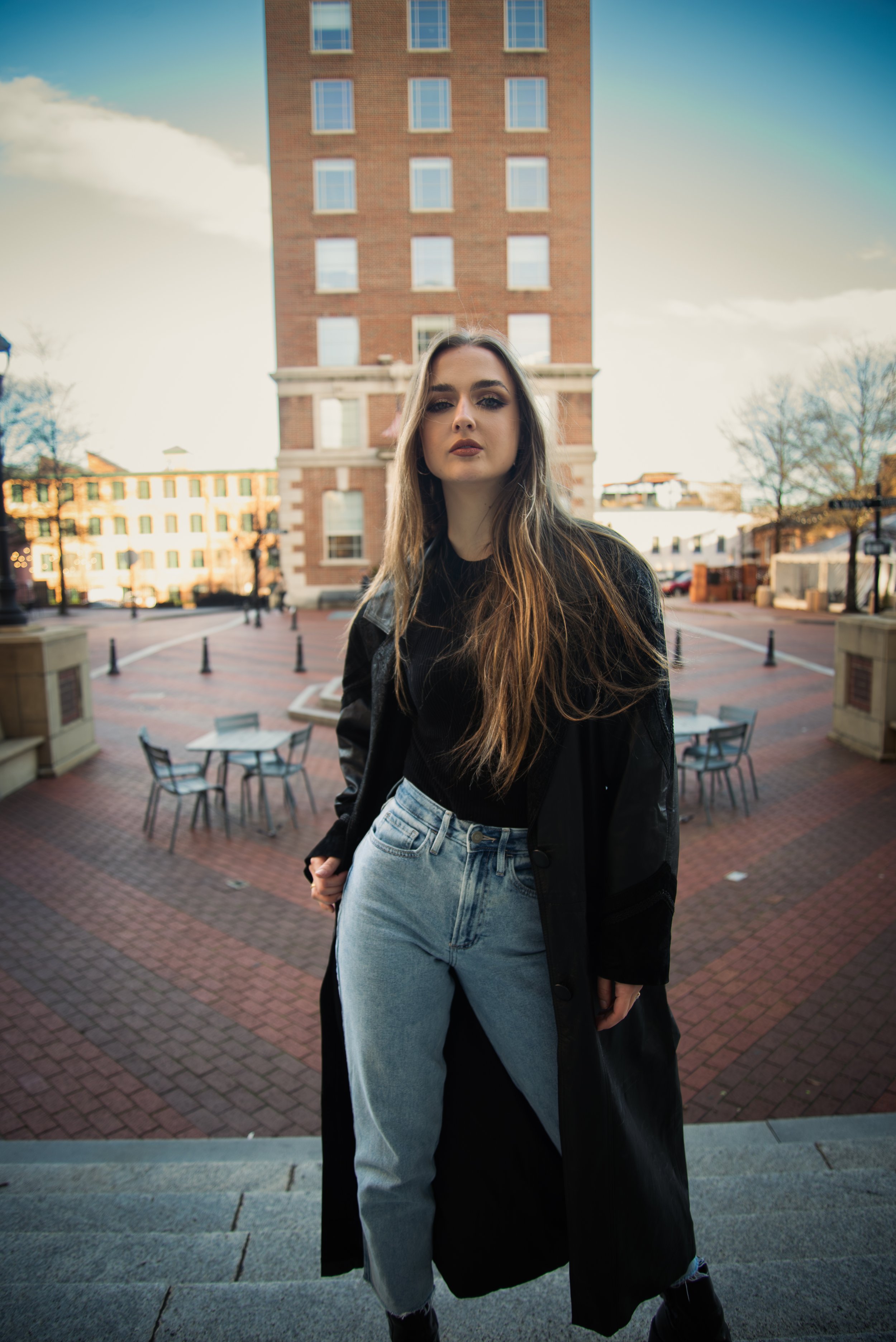 A young woman with long brown hair stands on steps outside in an urban area, with a tall brick building behind her and a blue sky.