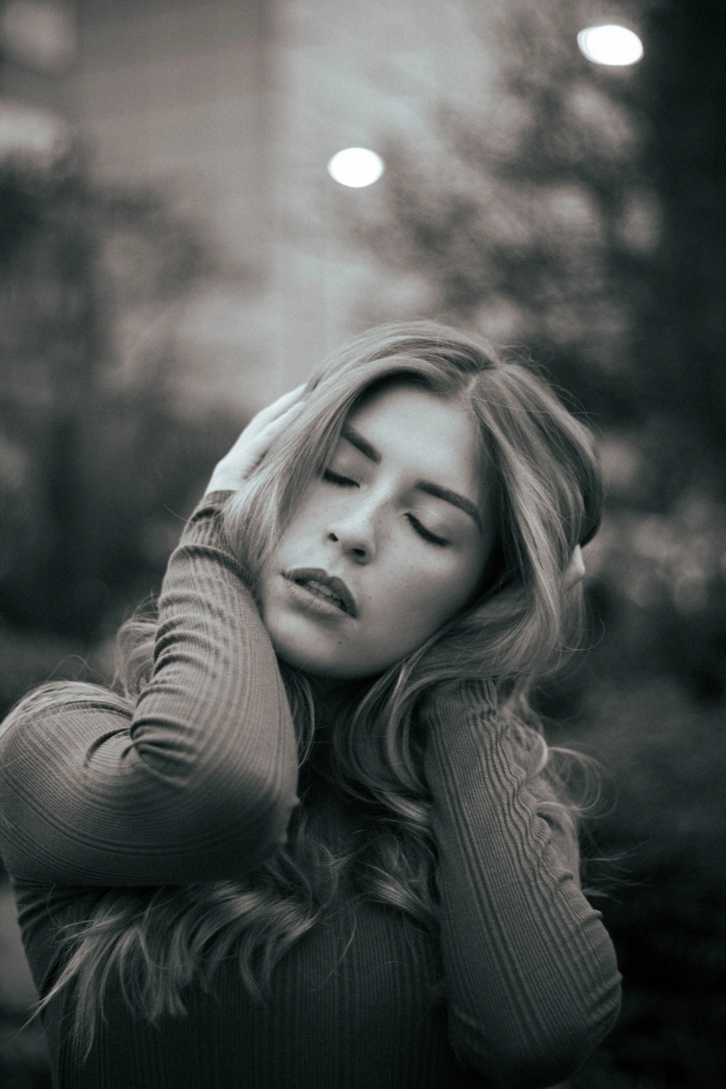 A woman with long, wavy hair holding her head with both hands, with her eyes closed and a serene expression on her face. The background is blurred with bokeh lights.