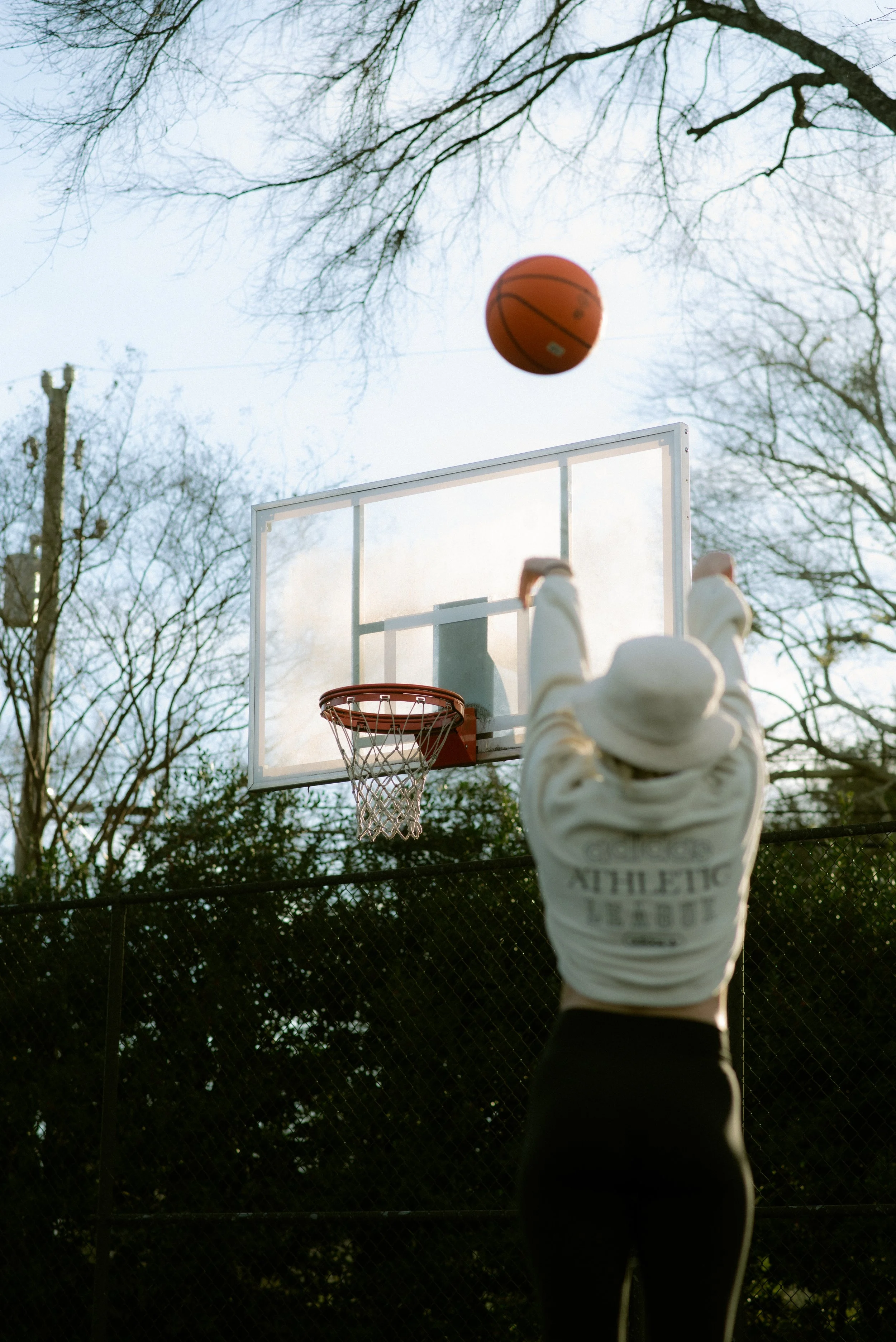 Person shooting a basketball towards the hoop on an outdoor basketball court, with bare trees in the background.