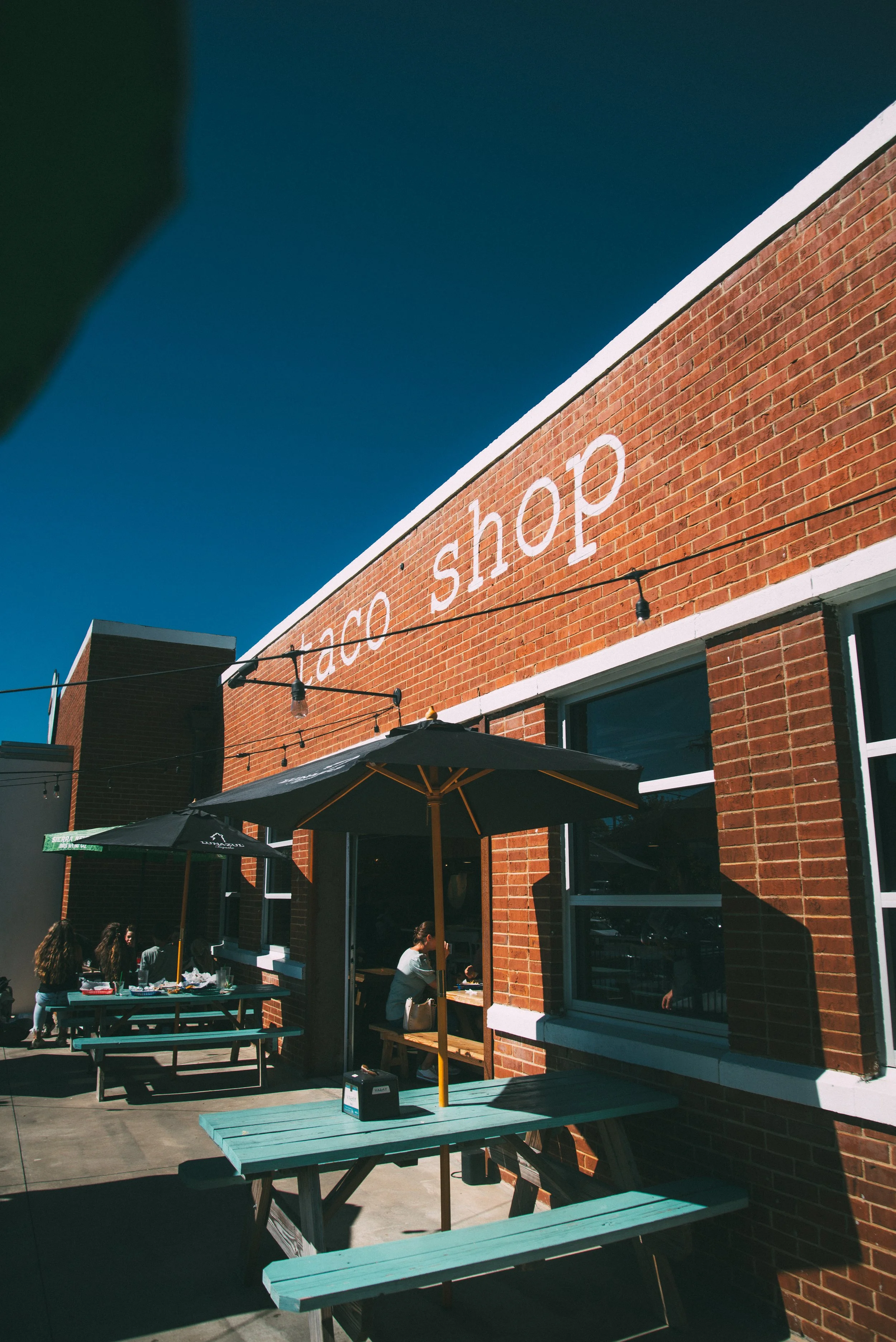 A brick building with a sign reading 'taco shop,' outdoor seating with Capri-style umbrellas, and people sitting and standing outside on a sunny day.