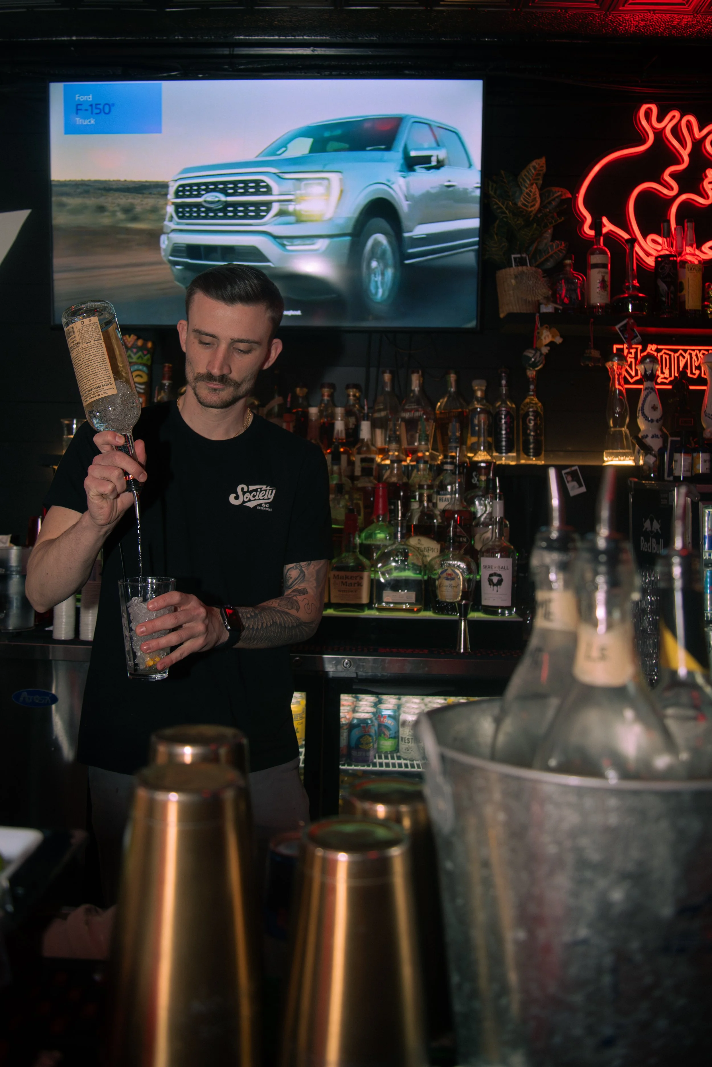A bartender with tattoos and a mustache is preparing a drink in a bar. There are various bottles of liquor on shelves, a large screen showing a blue Ford F-150 truck, and neon lights in the background.