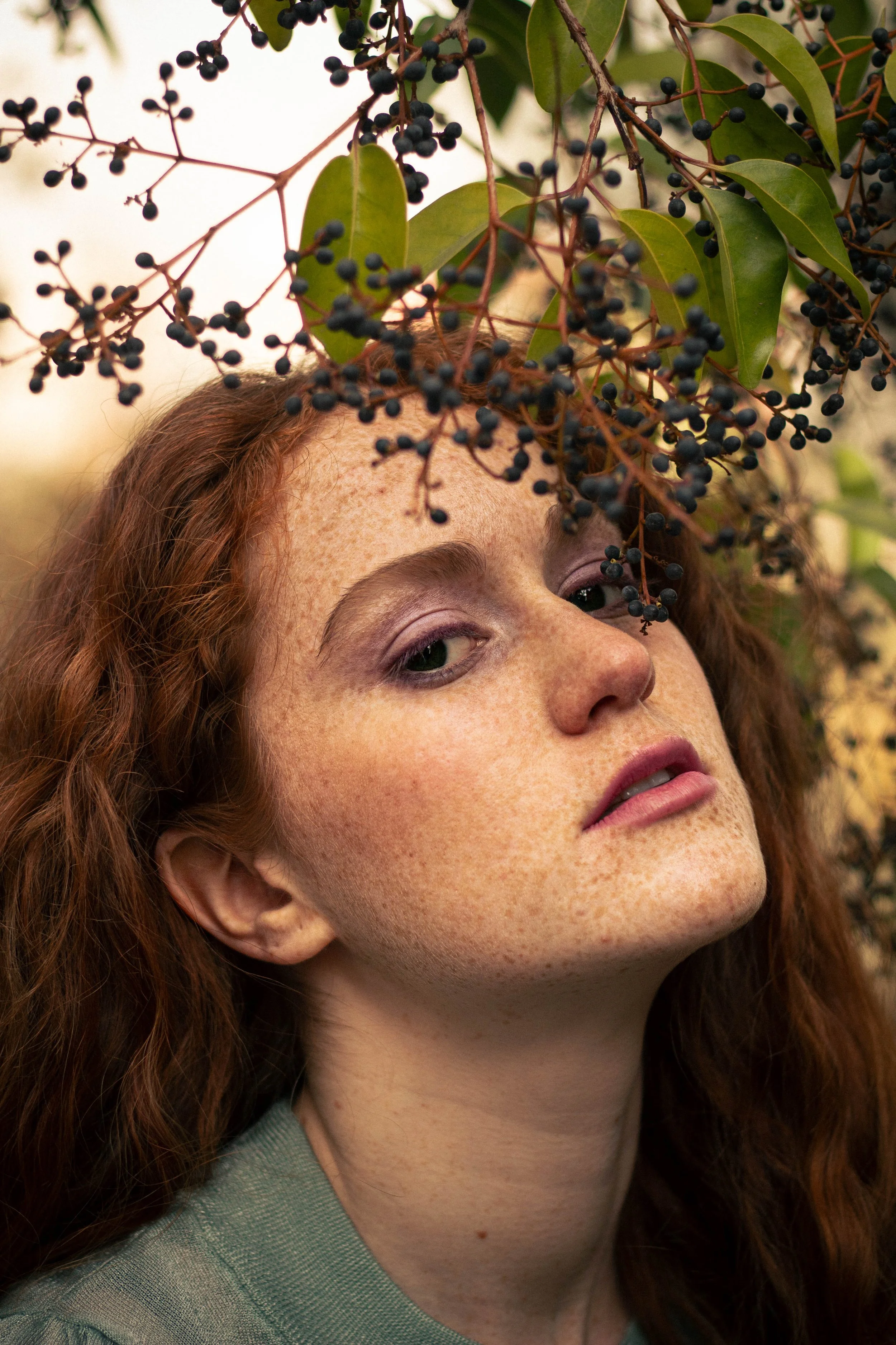 Close-up of a woman with red curly hair, freckles, and green eyes, partially obscured by branches with dark berries and green leaves.