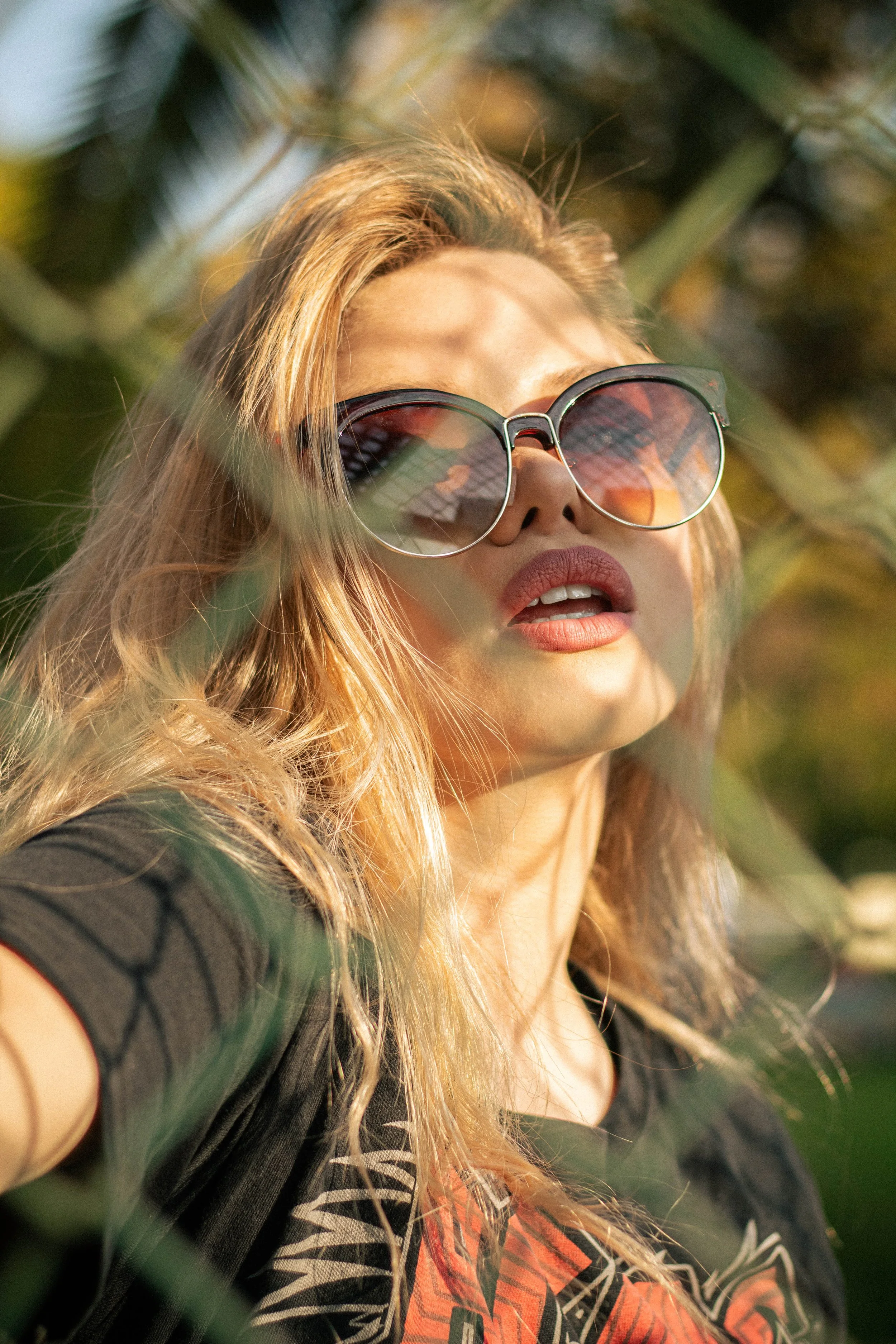 A woman with long blonde hair wearing sunglasses and a dark t-shirt with a graphic design. She is outdoors, partially obscured by green plants.