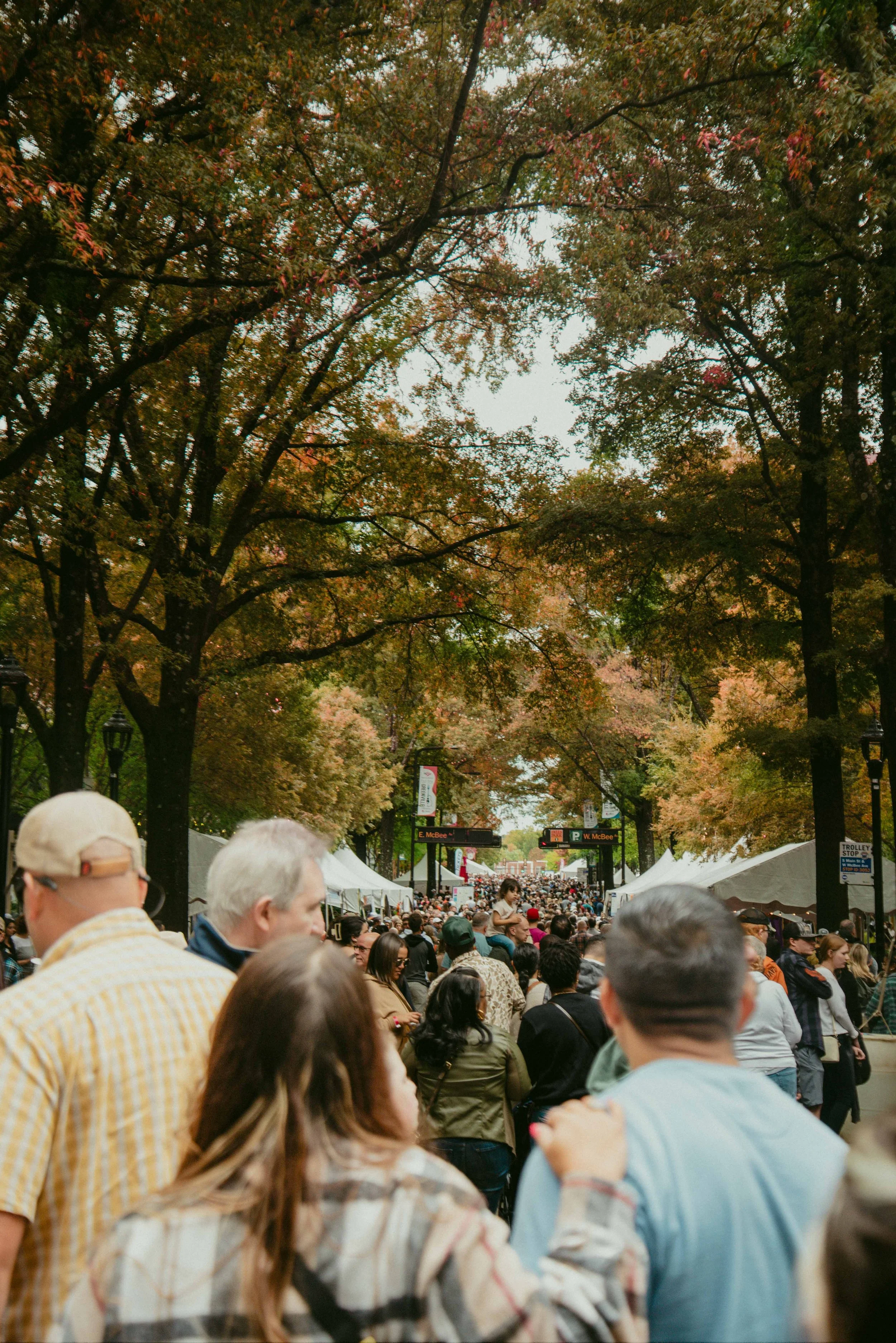 Crowd of people walking outdoors among large trees with colorful leaves on a fall day. Tents are set up along the sides of the walkway, with signs indicating street names.