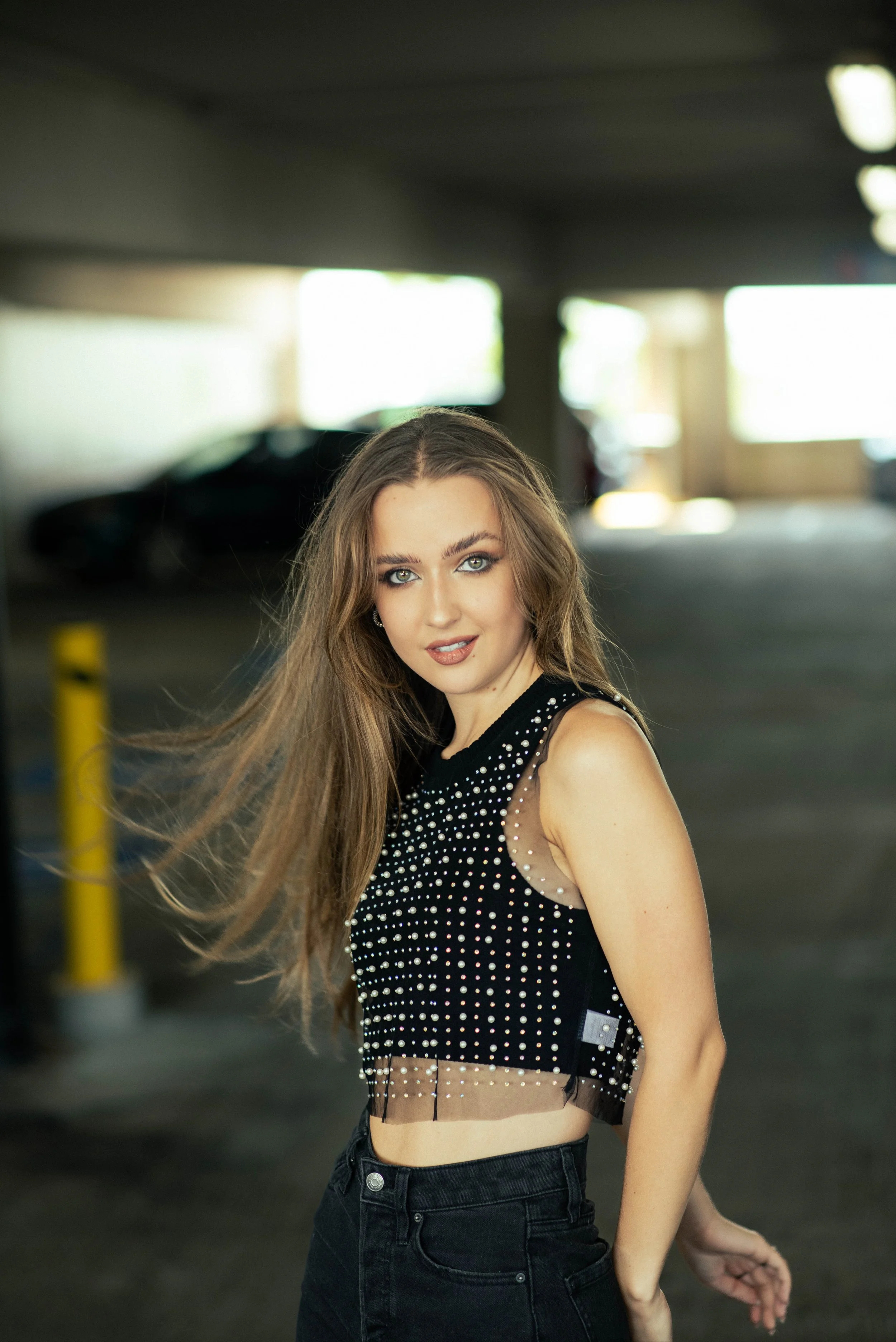 A young woman with long, wavy brown hair and light makeup, wearing a sleeveless black top decorated with small white pearls, standing in an indoor parking garage.