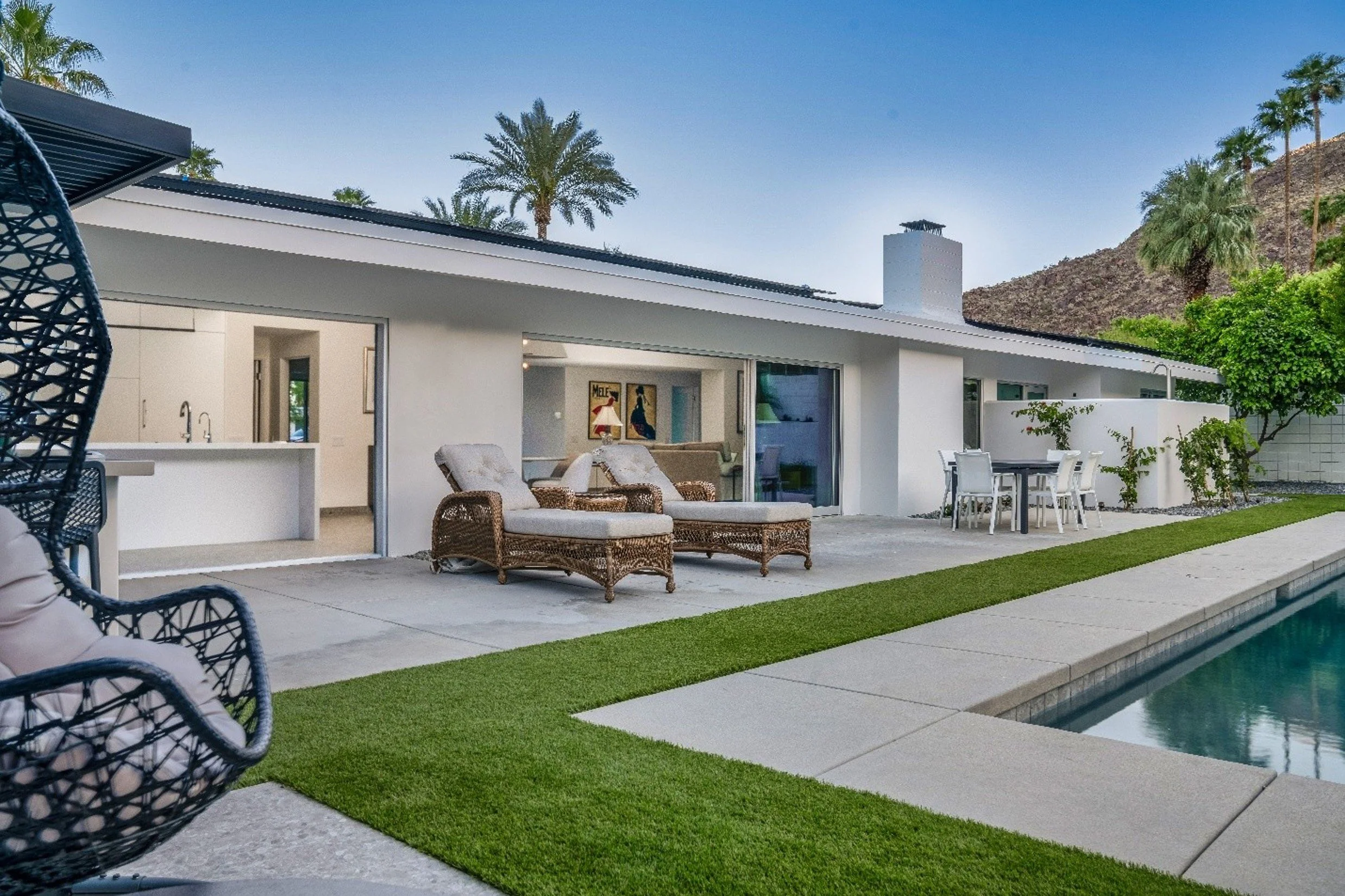 Backyard with swimming pool, outdoor lounge chairs, dining table with chairs, and a modern white house with large glass sliding doors, surrounded by palm trees and desert mountains in the background.