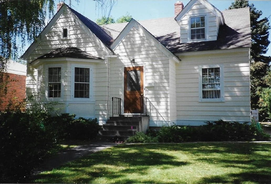 A two-story white house with a front door, three windows, and a small dormer window on the roof, surrounded by a lawn and trees.