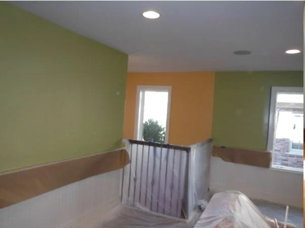 Room with green and orange walls, windows, and temporary plastic protective covering on staircase railing and furniture, indicating ongoing renovation or painting.