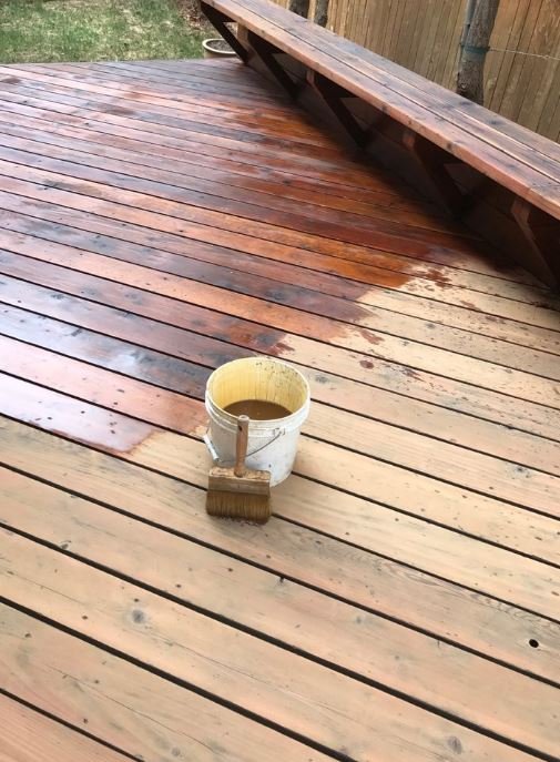 A wooden deck with a paint bucket and brush in the center, showing partially finished wood stain.