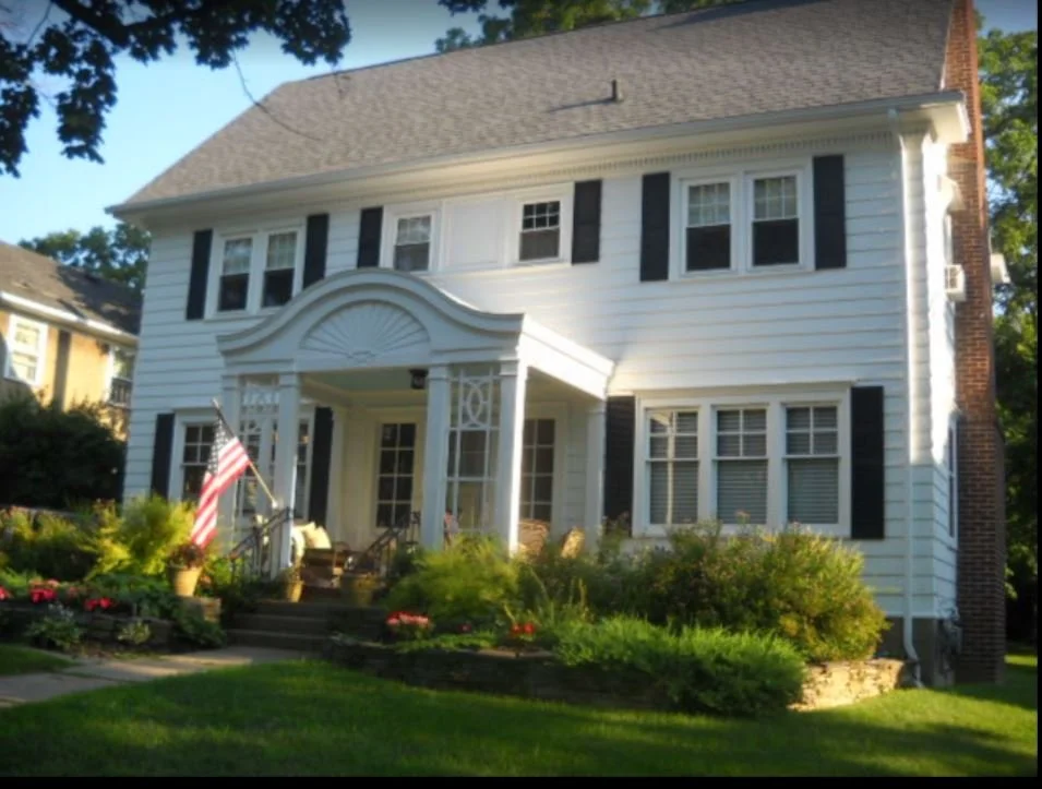 A white, two-story house with black shutters and a brick chimney, surrounded by green shrubs and a lawn, with an American flag near the front porch.