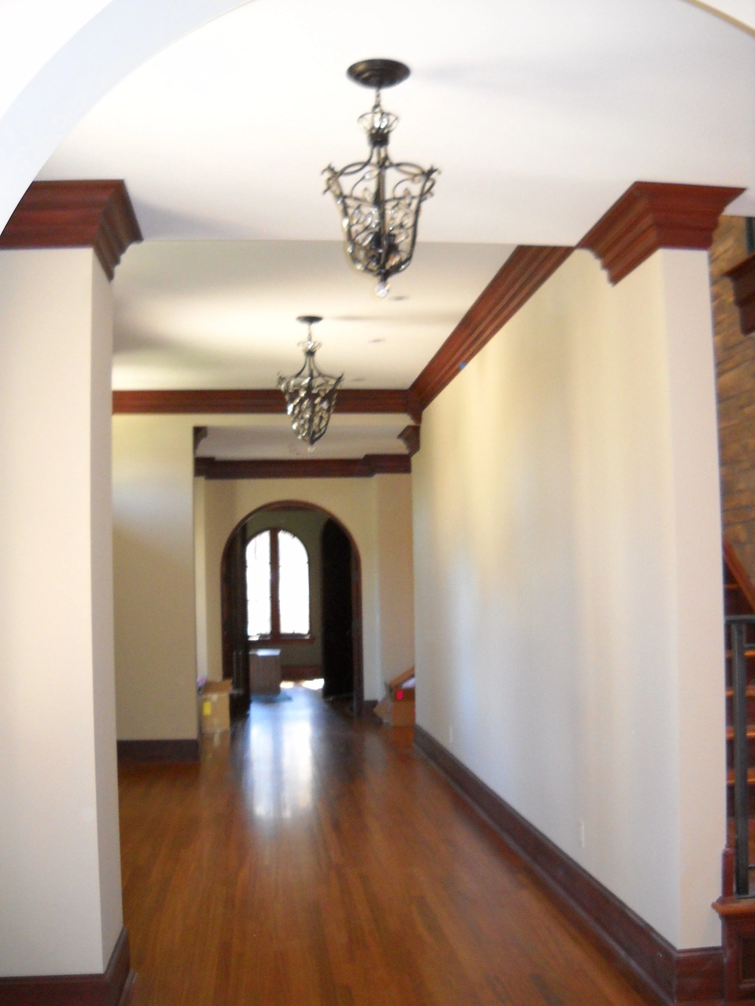 Interior view of a hallway with hardwood floors, white walls, wooden crown molding, and two decorative chandeliers hanging from the ceiling.