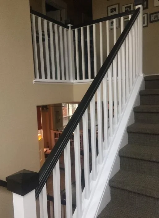 Indoor staircase with white railing and gray carpeted steps, leading up to a second floor. A black handrail runs along the white balusters. Part of a wall with picture frames is visible.