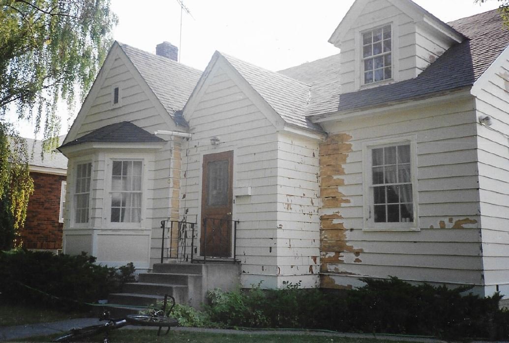 A white house with peeling paint on the exterior wall, front porch with stairs, and multiple windows. The house shows signs of deterioration.