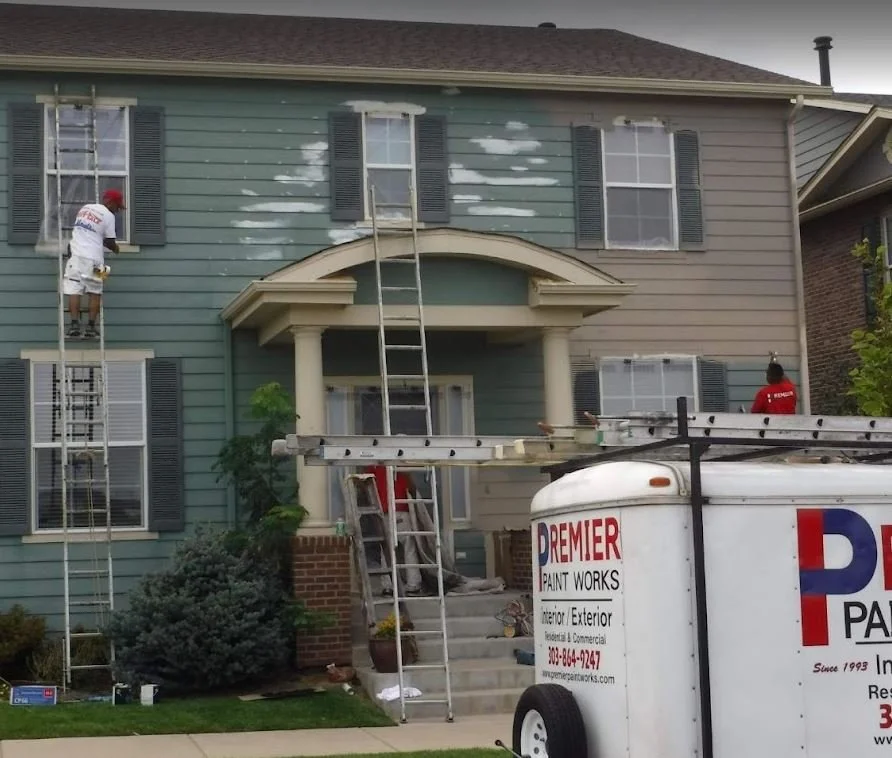 Two painters working on the exterior of a two-story house, one on a ladder and one on a scaffold, painting the upper walls in a different color. A van from Premier Paint Works is parked in front.