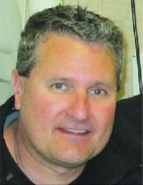 A close-up photo of a smiling man with short, curly gray hair, wearing a black shirt, indoors.