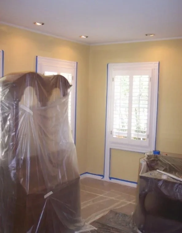 A room being prepared for painting or remodeling, with furniture and windows covered in plastic, and painter's tape applied around the walls and floor.
