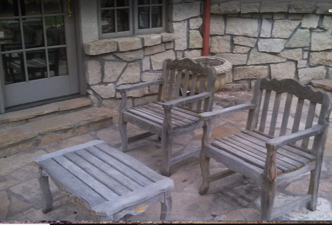 Three wooden patio chairs and a footstool in front of a stone building with a glass door and window, and a large decorative stone planter behind the chairs.
