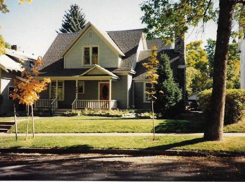 A two-story gray house with a front porch, surrounded by trees and a well-maintained lawn in a suburban neighborhood.