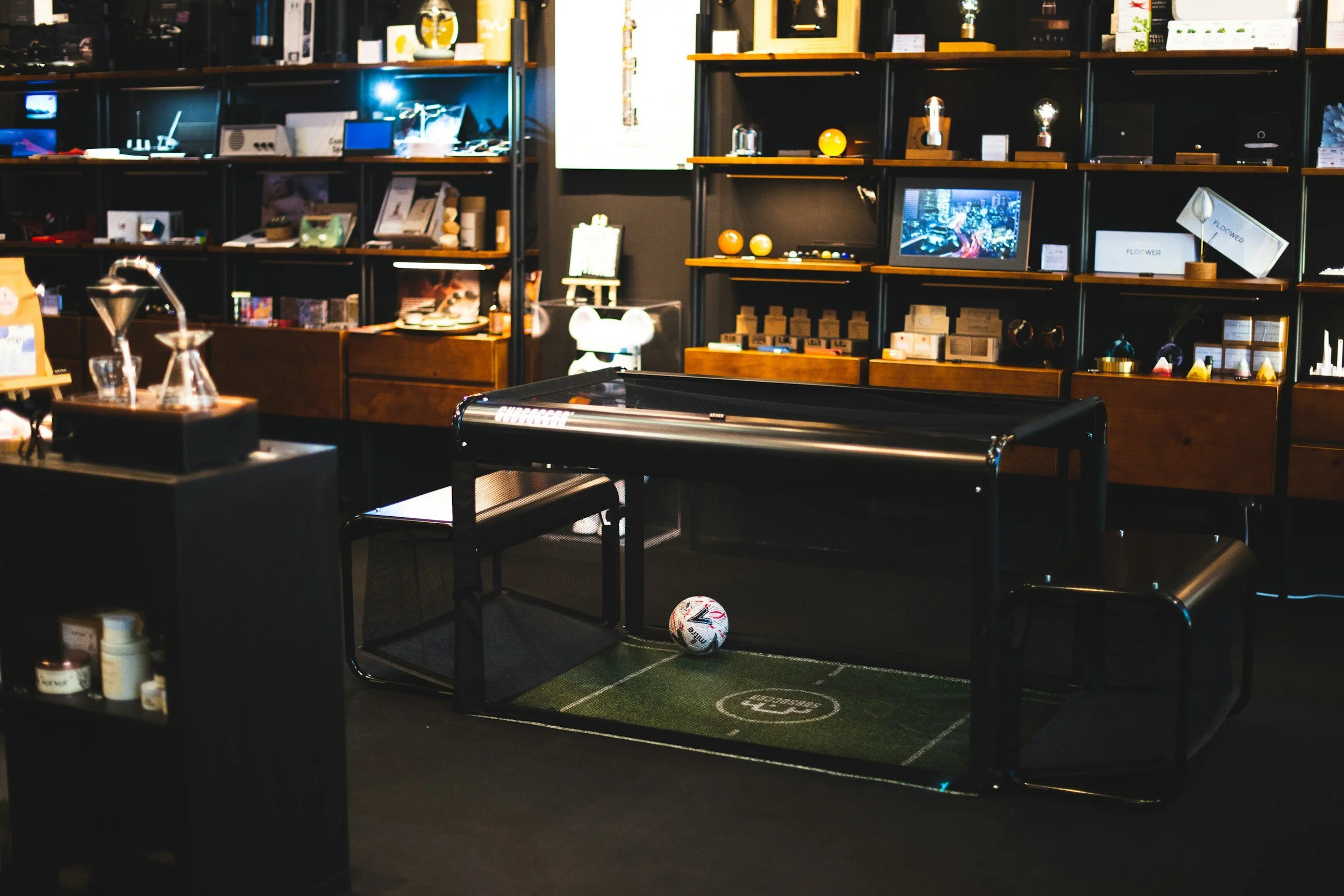 Indoor space featuring a small soccer field with a ball, a black cushioned bench, and shelves with various electronic gadgets, decor items, and framed pictures in the background.