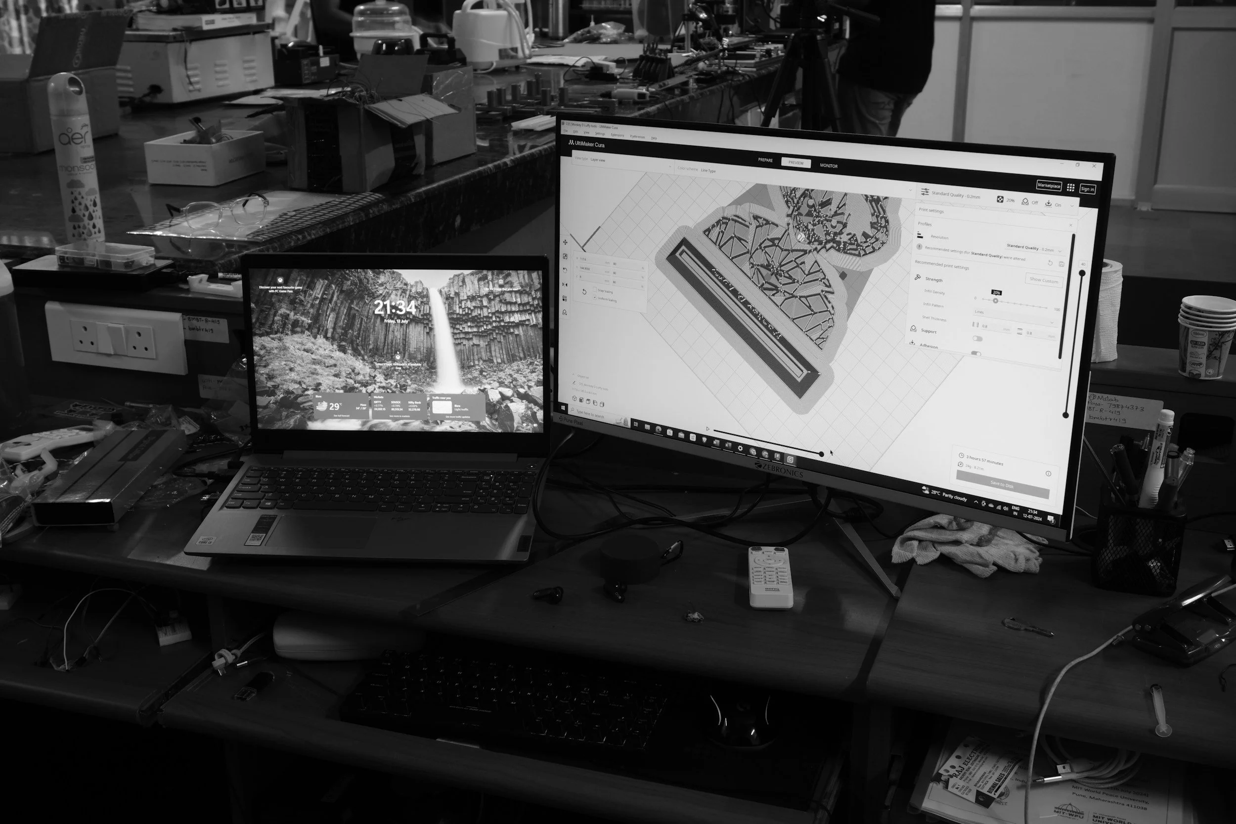 A cluttered workspace with a laptop and a large monitor displaying 3D modeling software, along with various objects on the desk including cables, a remote, and papers.