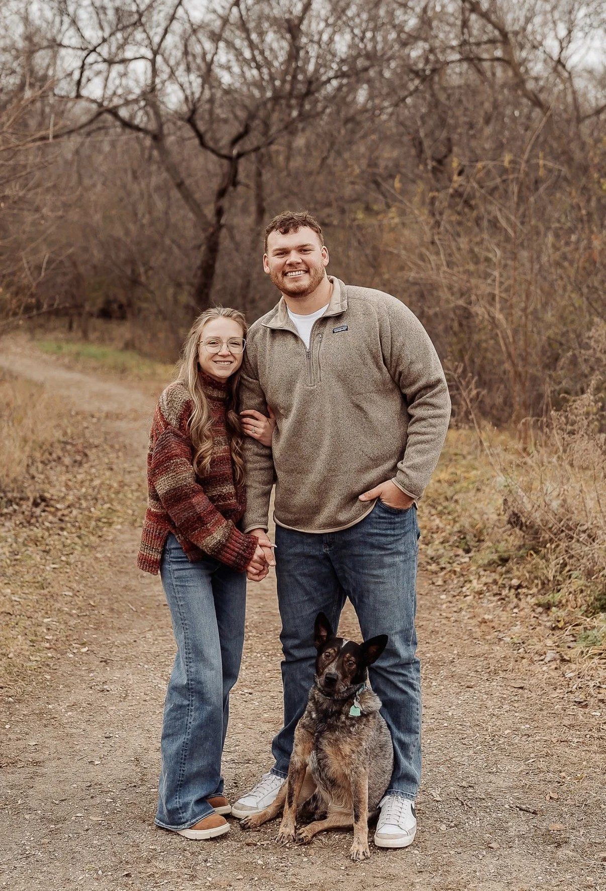 A smiling couple and their dog on a dirt path in a wooded area during fall. The woman has long wavy hair, glasses, and wears a striped sweater. The man has short curly hair, a beard, and wears a beige fleece jacket. The dog is sitting and has a brindle coat.
