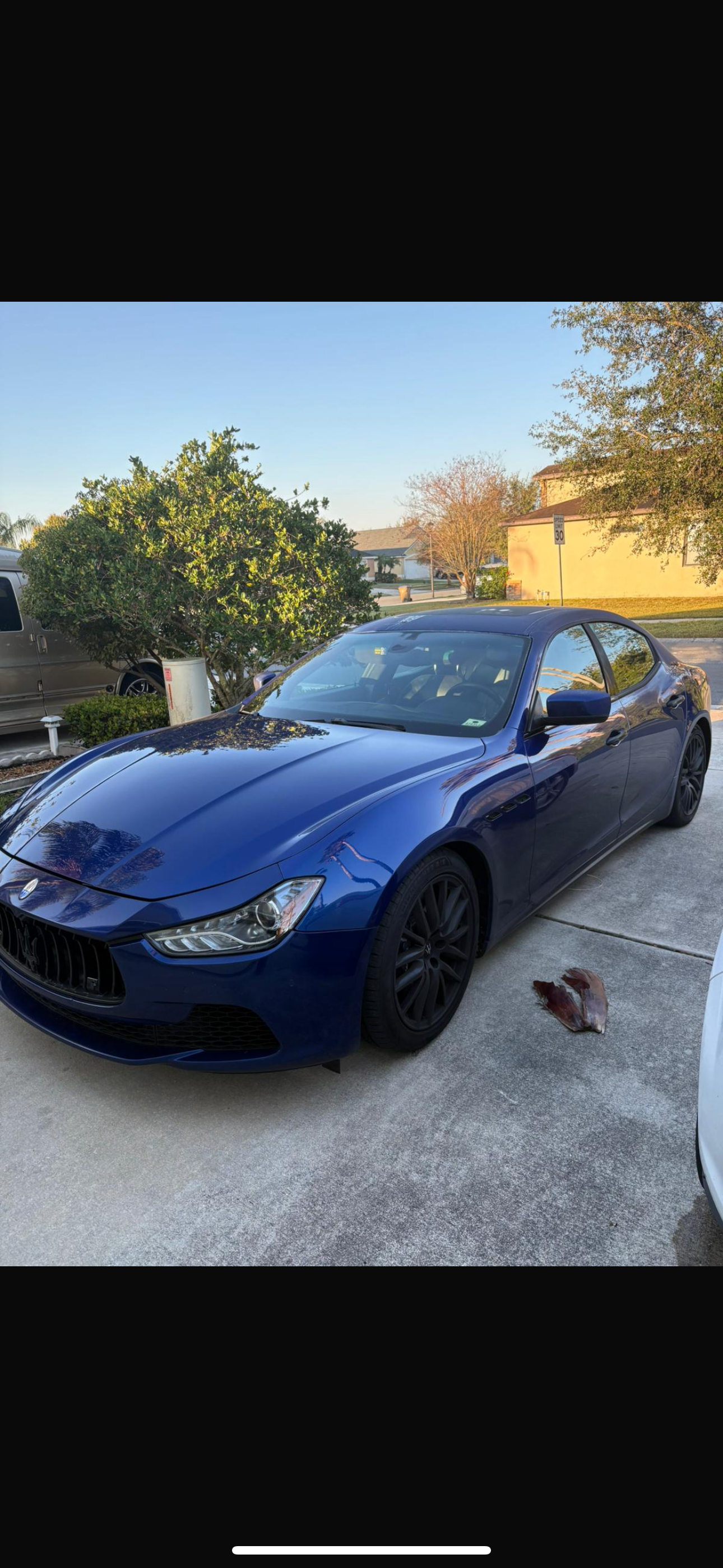 Blue sports car parked in driveway with a tree and house in the background, and a dried leaf on the ground near the car.