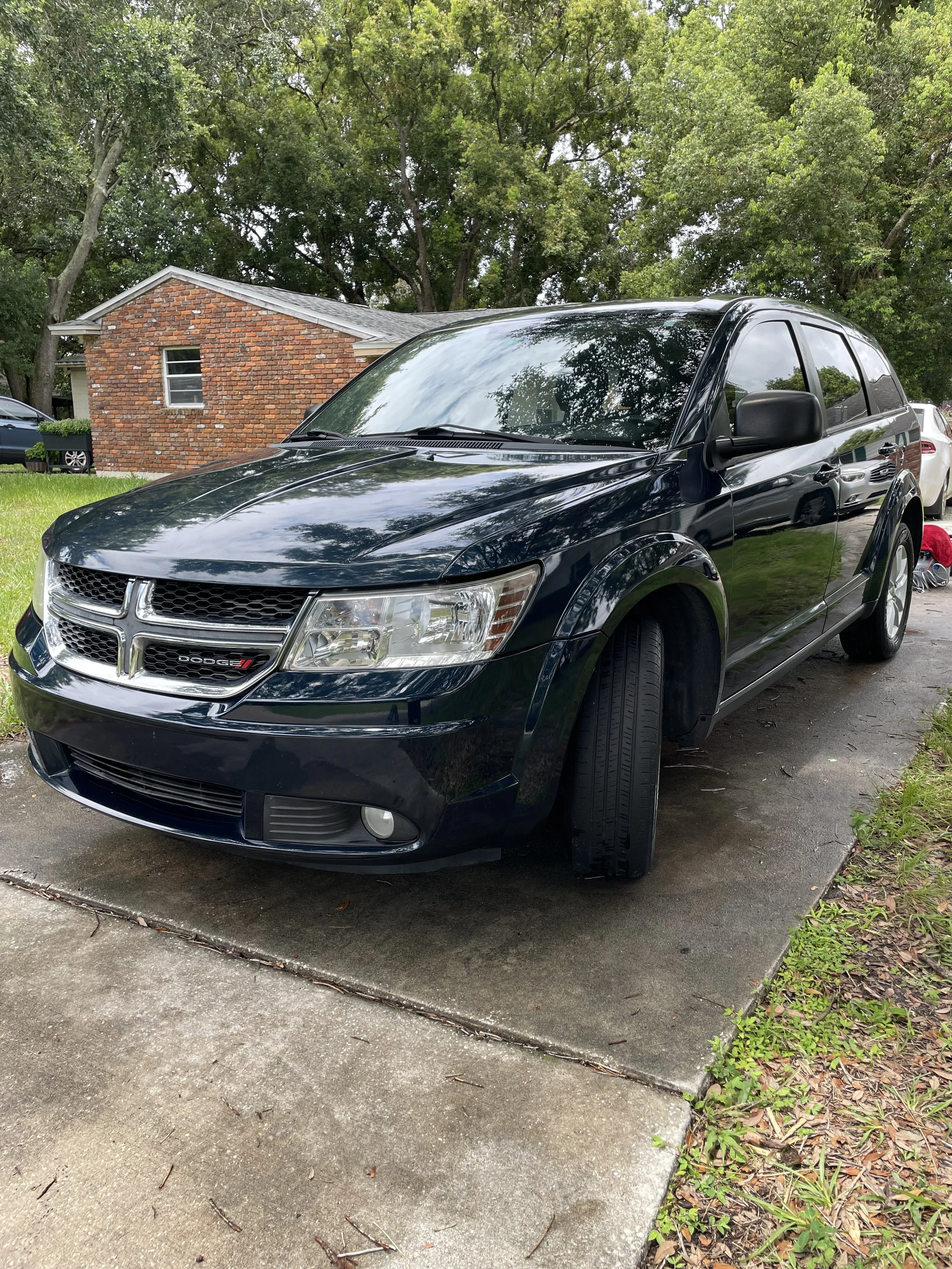 Black Dodge SUV parked on driveway with missing front right tire, green trees in background, house with brick exterior behind.
