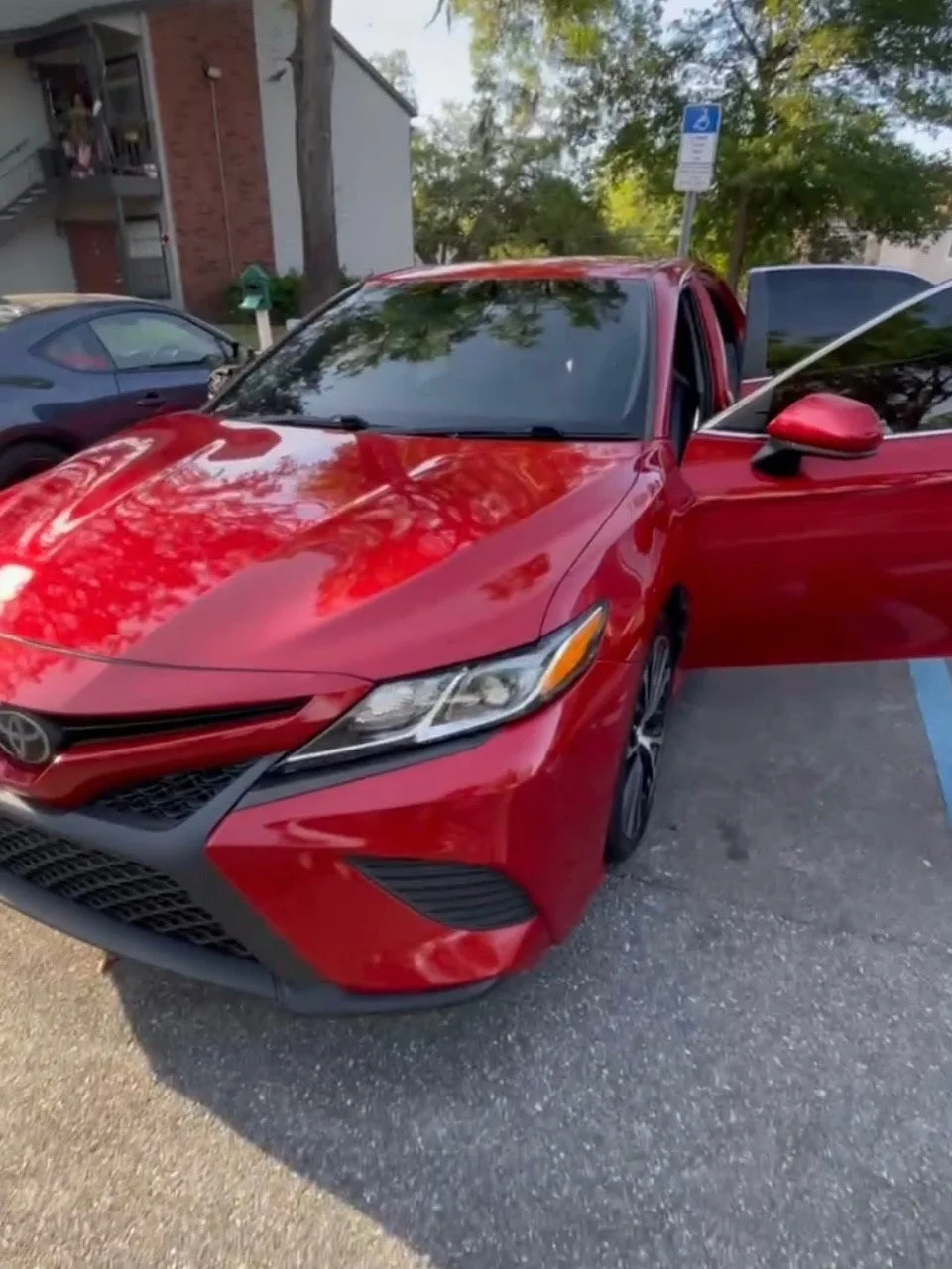 Red sedan parked in a handicapped spot with its front open, in a parking lot near a building and trees.