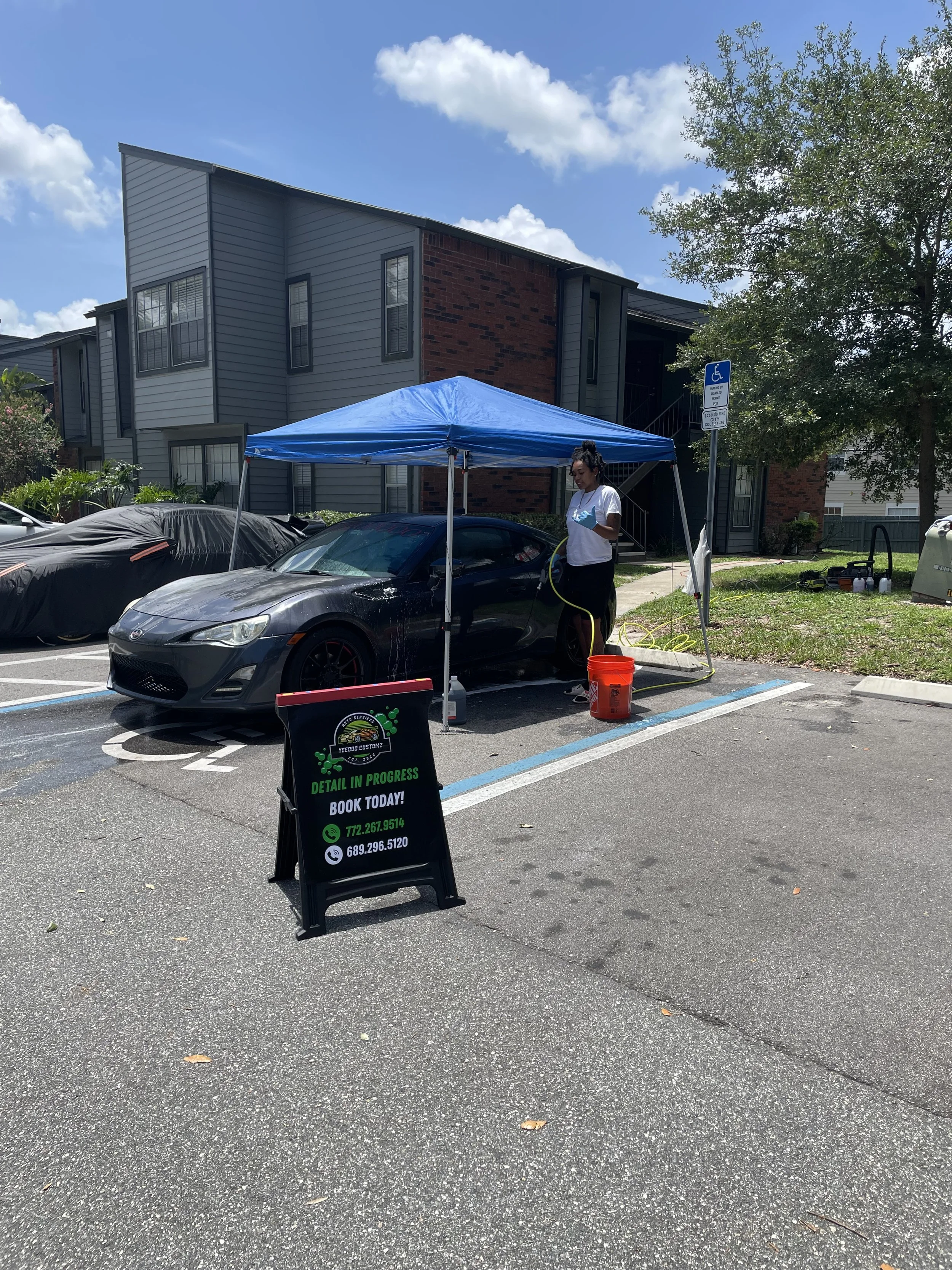 A person is washing a black sports car under a blue canopy in a parking lot, with a sign advertising detailing services and contact information.