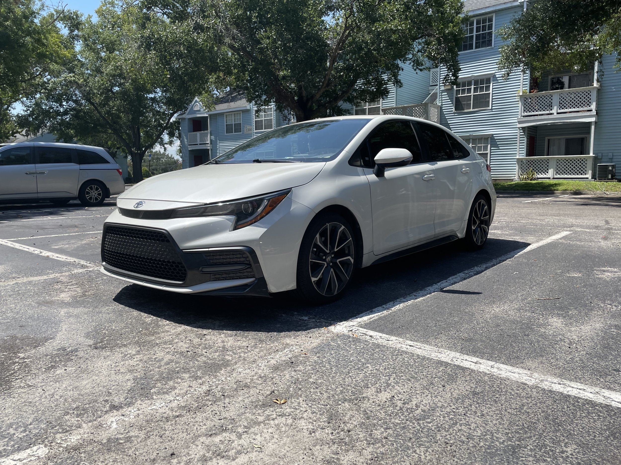 A white Toyota Corolla parked in an outdoor parking lot during daytime, with other vehicles and residential buildings in the background.