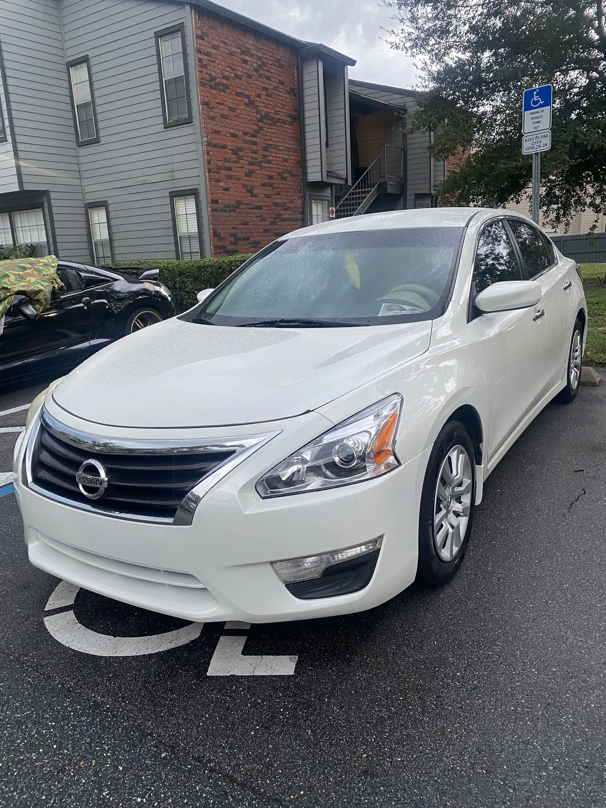 A white Nissan sedan parked diagonally in a disabled parking space on a rainy day, with water droplets on the car and a building in the background.
