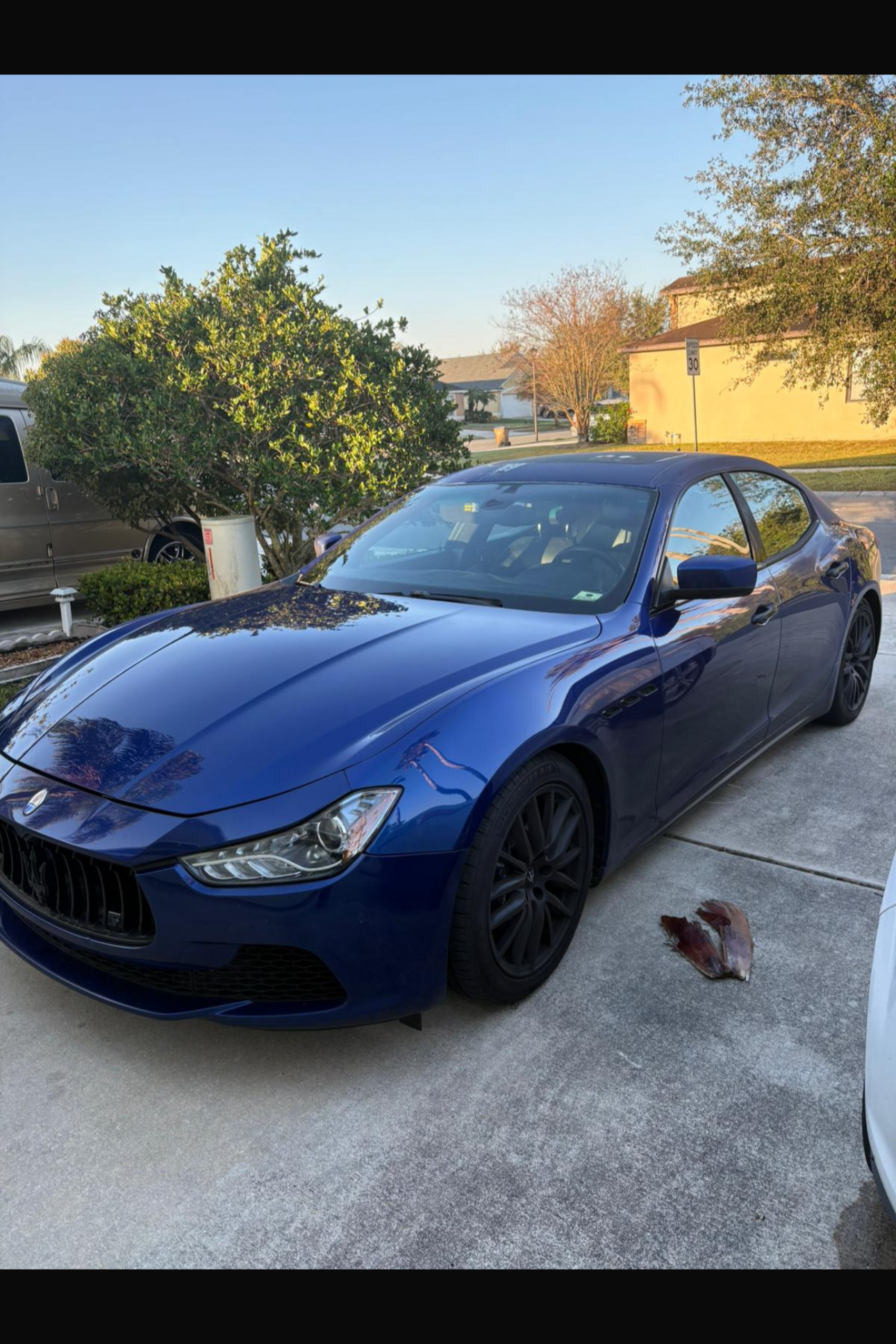 A blue Maserati parked on a residential driveway with a piece of wood or bark on the ground nearby. The background shows trees, a house, and a street sign indicating a speed limit of 30 mph.