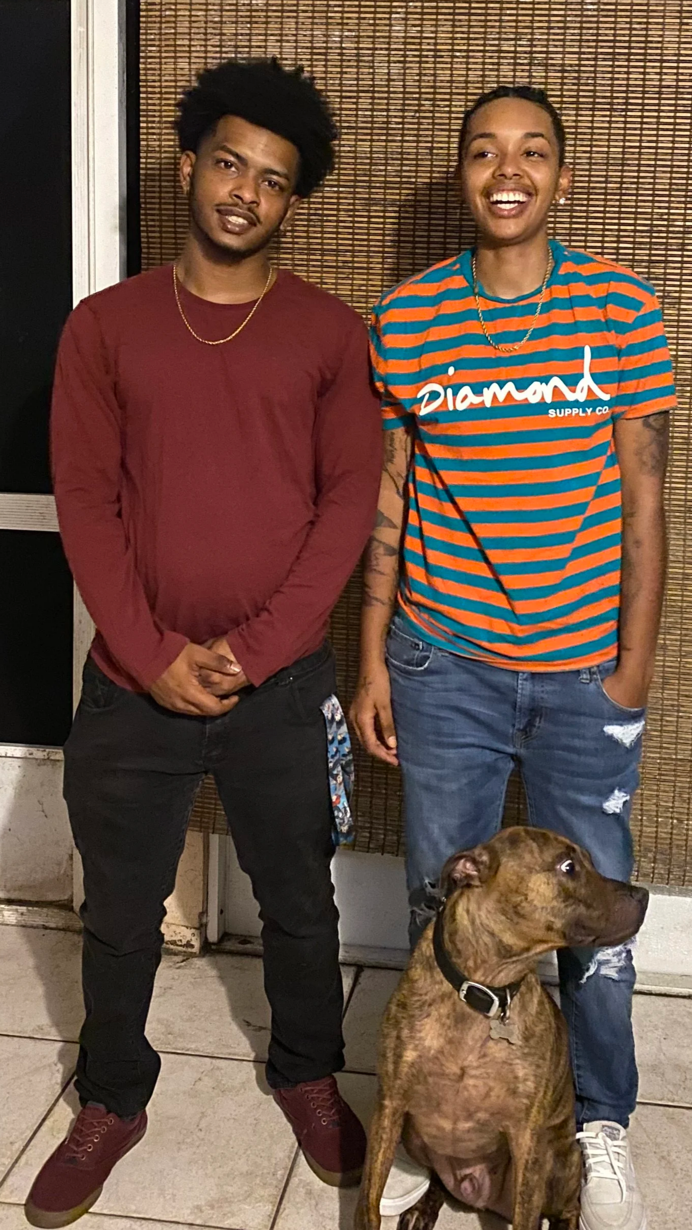 Two young men standing next to each other in front of a woven bamboo background with a dog sitting at their feet. The man on the left is wearing a maroon long-sleeve shirt, black distressed jeans, and maroon shoes. The man on the right is wearing a blue and orange striped T-shirt with the word 'diamond' on it, ripped jeans, and white sneakers. Both are smiling, and the dog is brown with a brindle pattern, sitting and looking to the side.