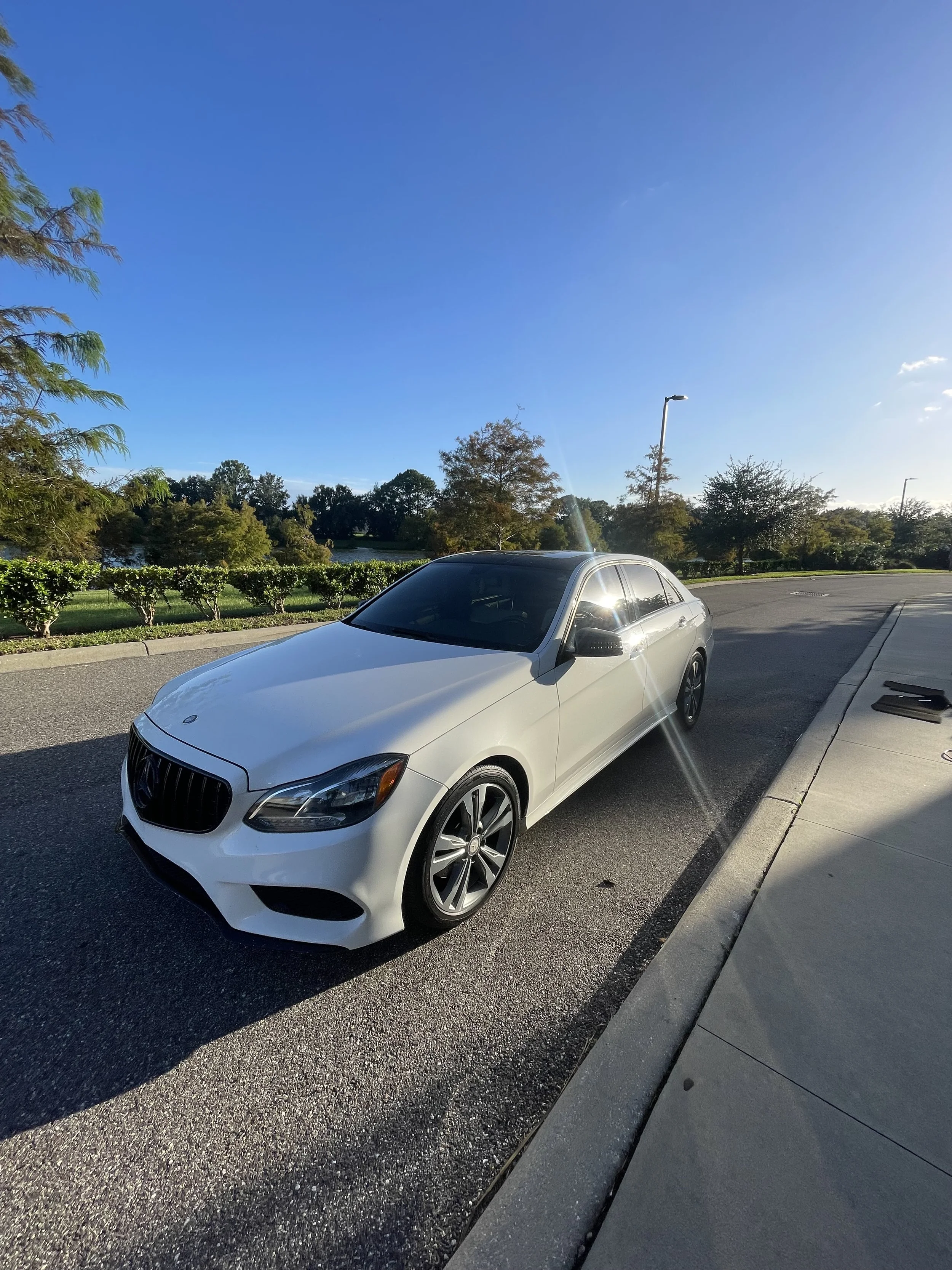 A white Mercedes-Benz sedan parked on the side of a paved road with green trees and a blue sky in the background.