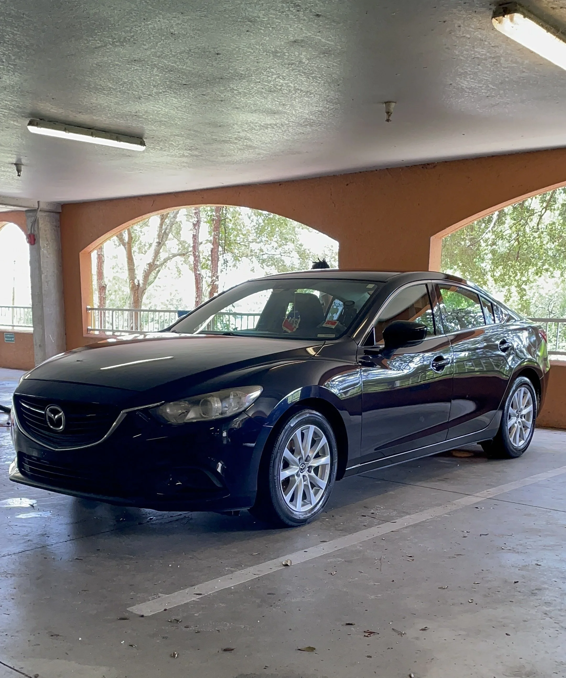 A black Mazda sedan parked in a covered parking lot with trees visible through large open windows in the background.