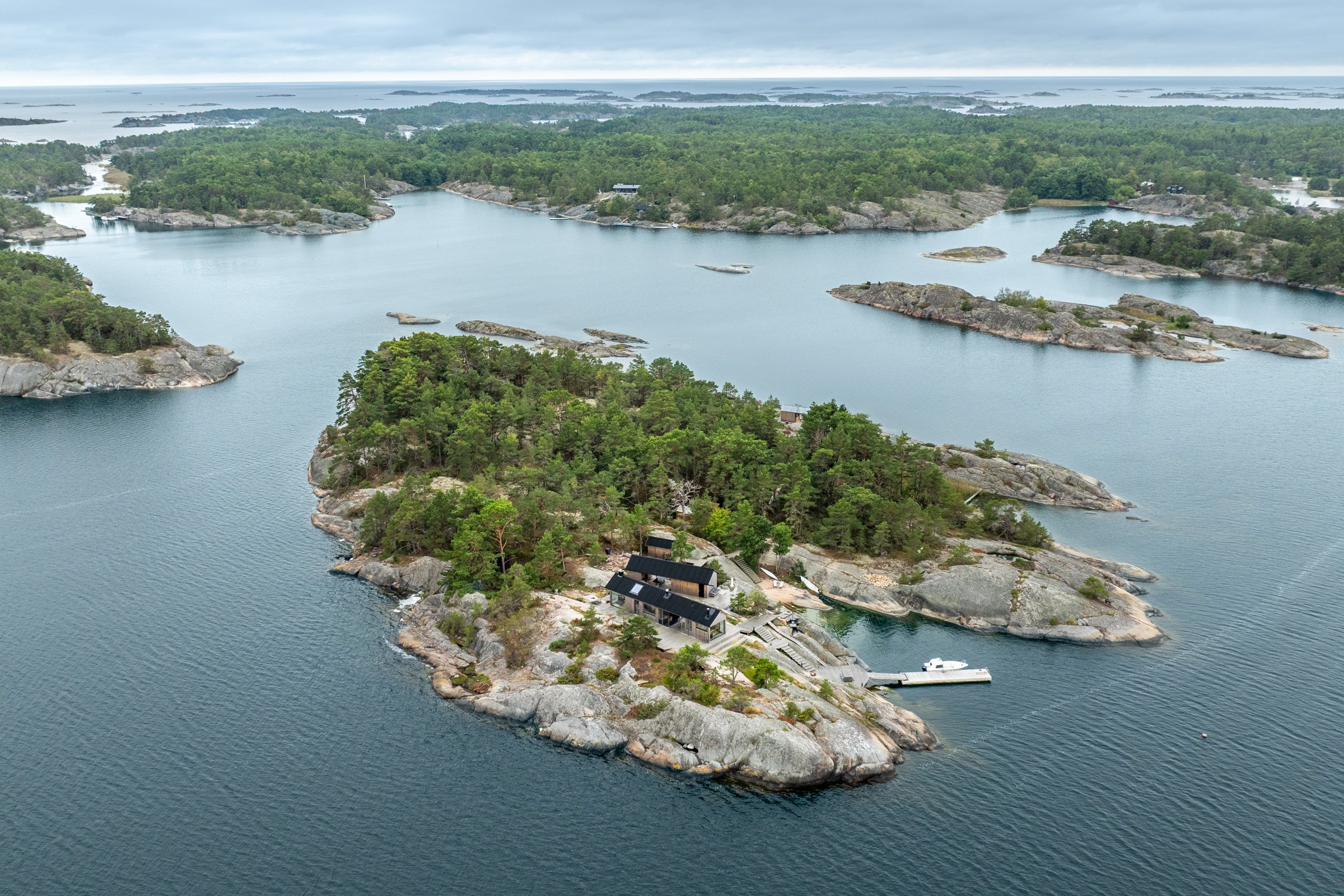 An aerial view of a small island with trees and rocky shores, featuring a house with a black roof and a dock with a boat, surrounded by calm water and other islands in the distance under an overcast sky.