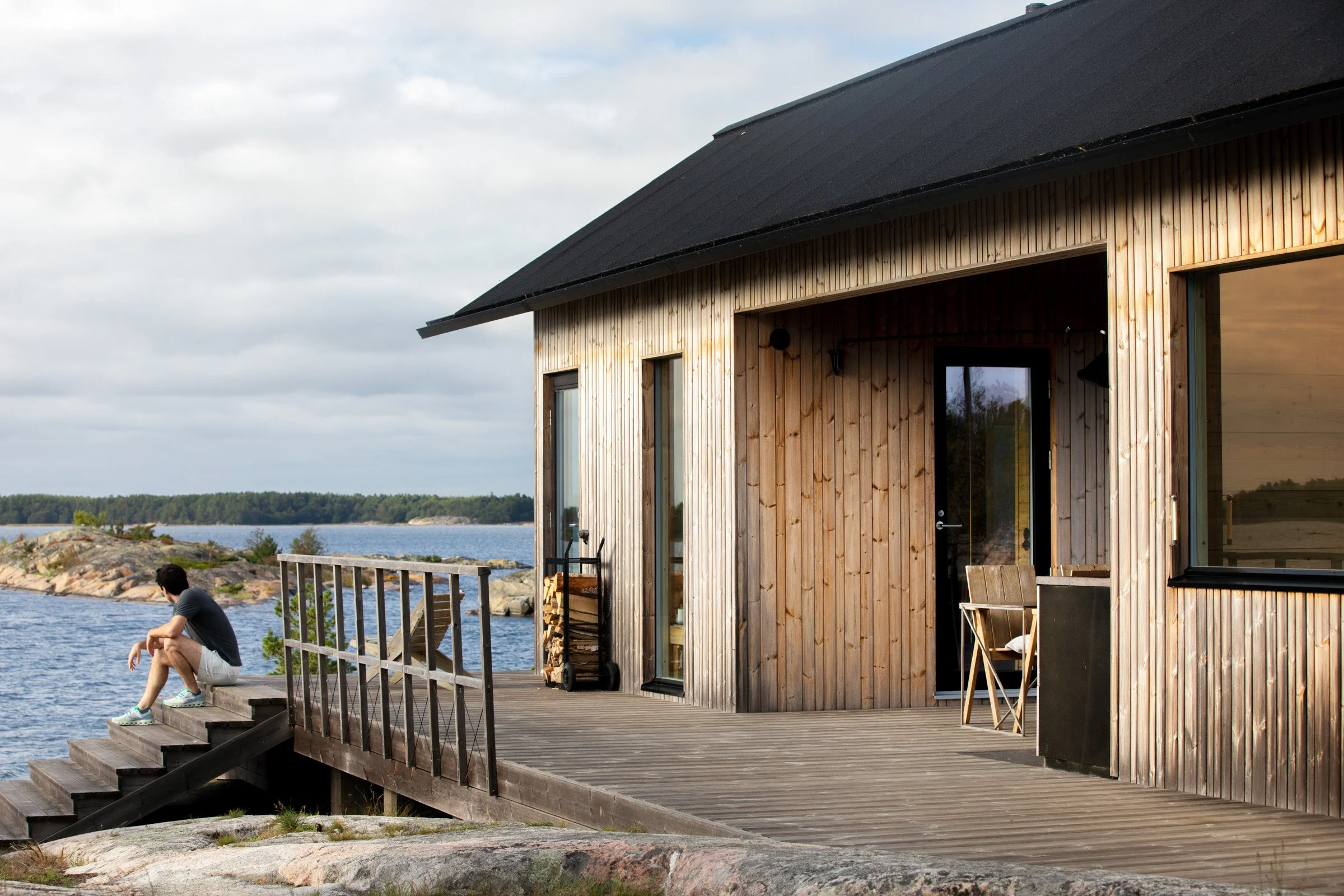 A person sitting on the steps of a wooden deck outside a modern wooden house by a lake, with water and rocks in the background, under a cloudy sky.