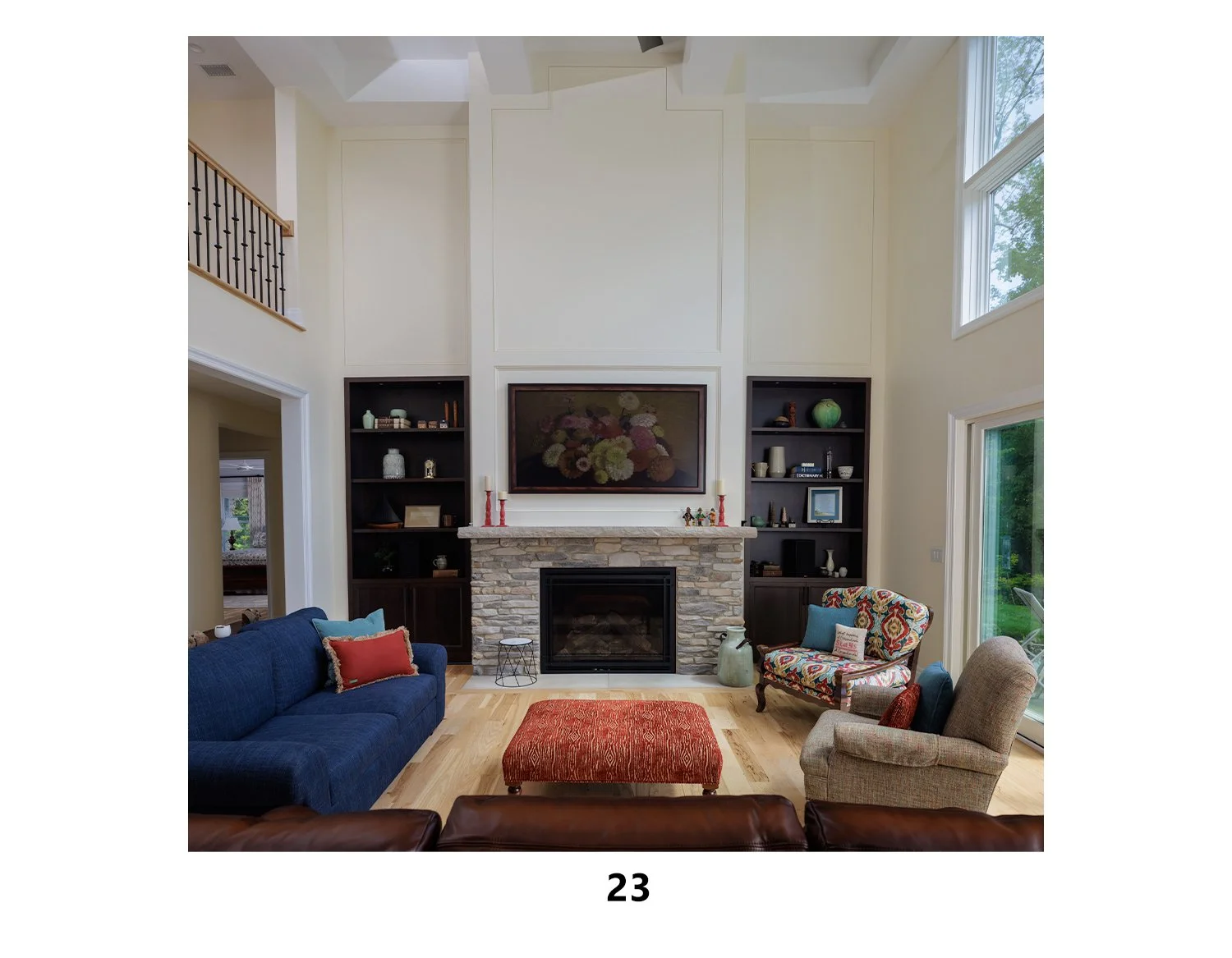 Living room with a stone fireplace, dark shelves on either side, colorful furniture including a blue sofa, patterned armchair, beige armchair, an orange ottoman, and decorative pillows, large windows, and hardwood flooring.