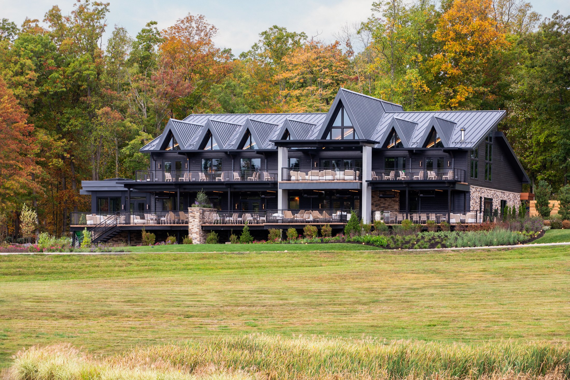 A modern multi-story house with black exterior and large windows, surrounded by trees with fall foliage, and situated on a well-maintained lawn with a garden.