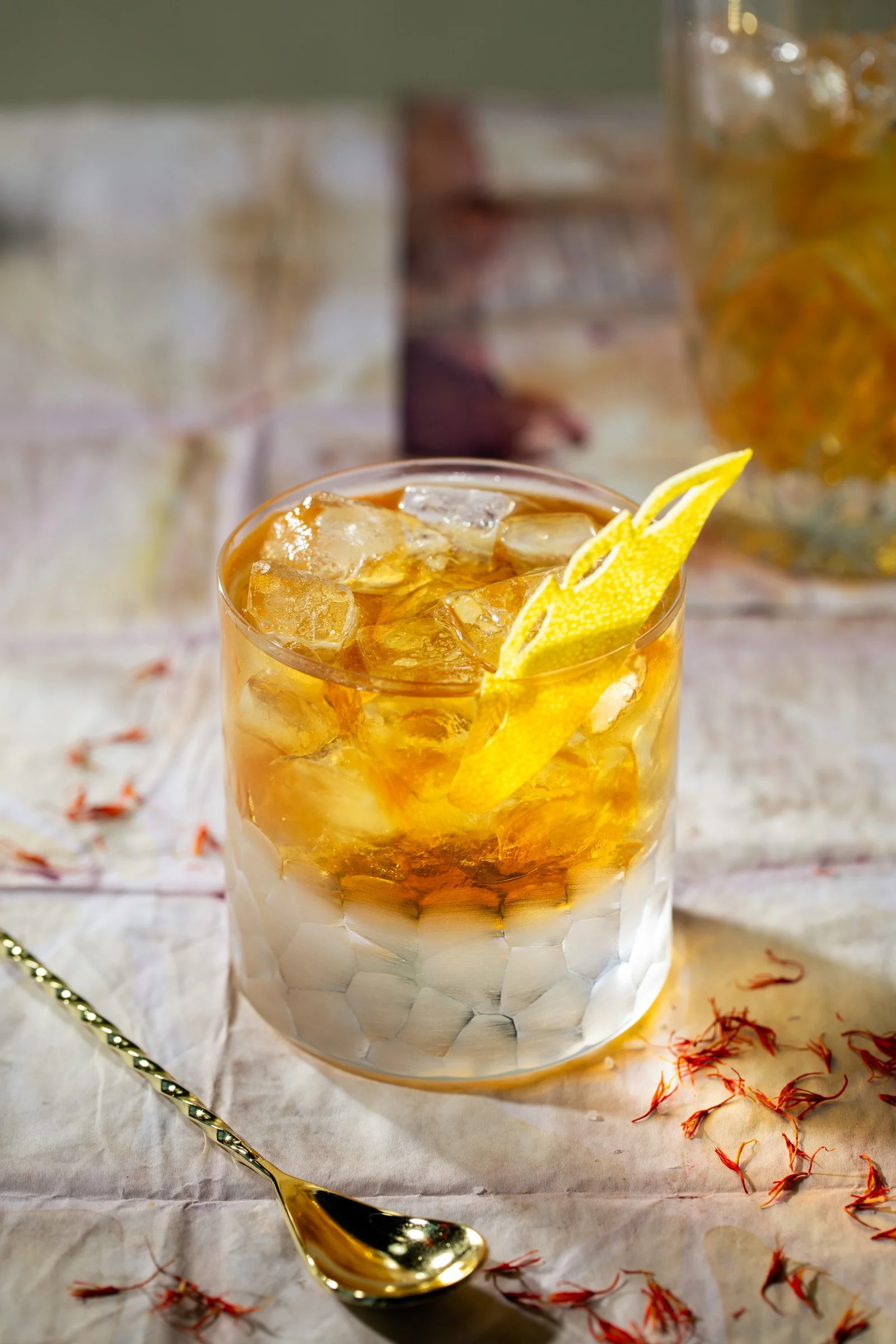 A glass of iced tea with lemon slices on a white cloth table, with a golden spoon and scattered saffron threads nearby.