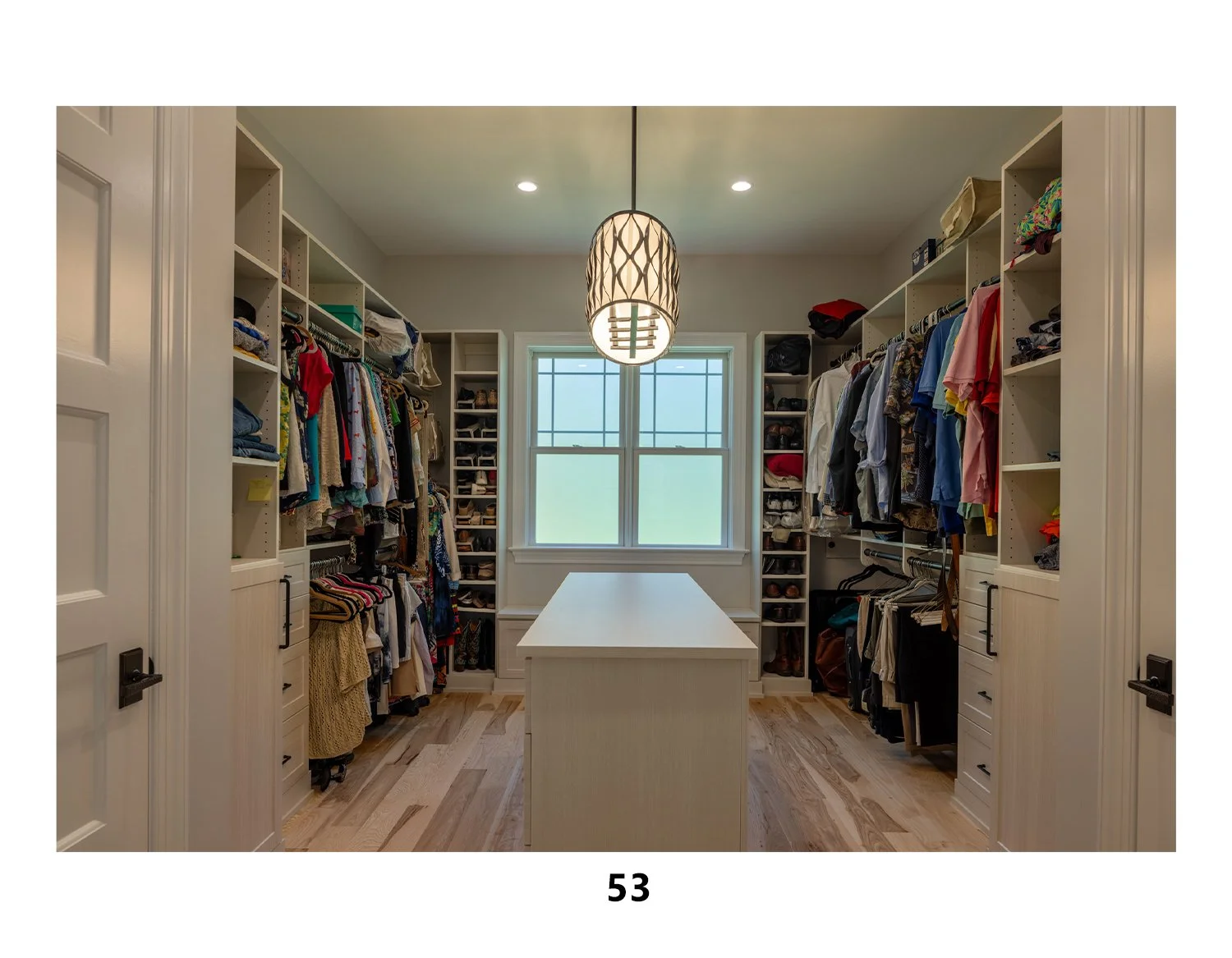 A walk-in closet with white built-in shelves filled with clothes, shoes, and accessories. A central white island. A window with natural light, ceiling lights, and a decorative pendant lamp.