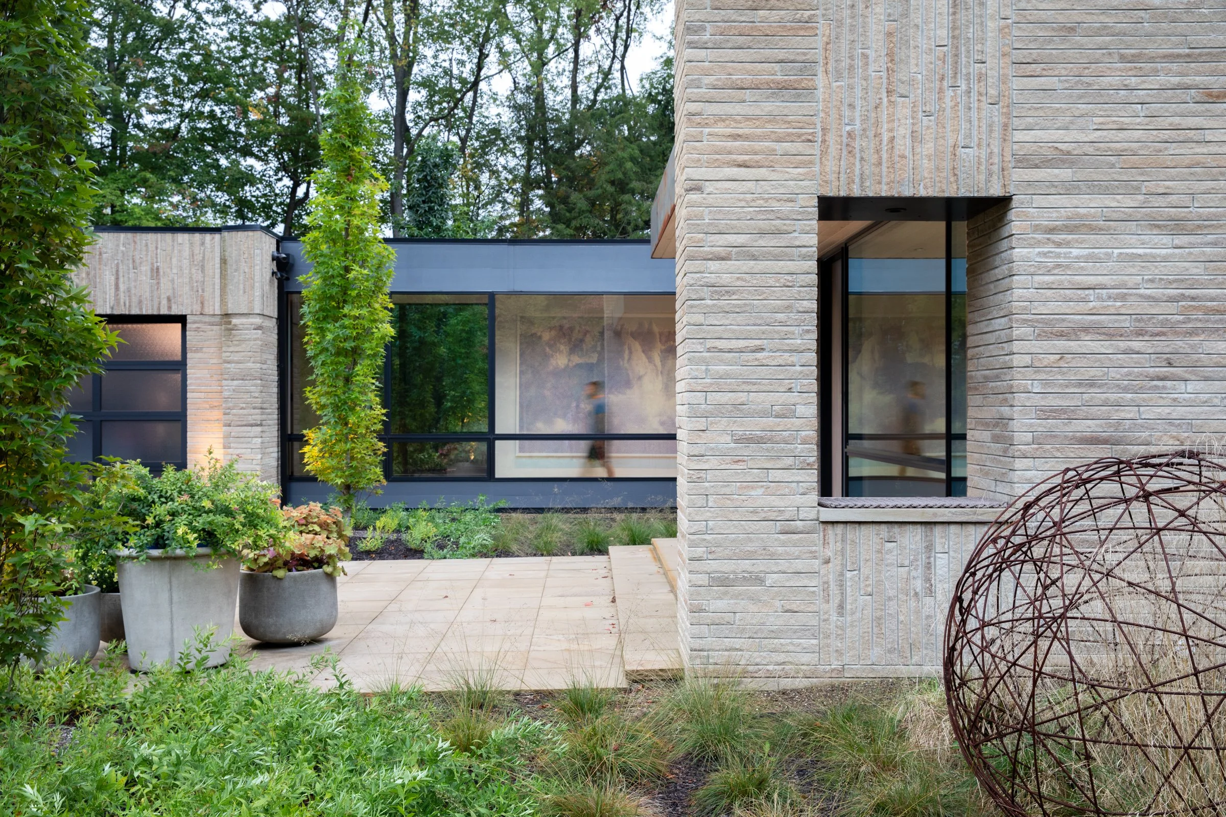 Modern house with beige brick exterior, black trim, large windows, and surrounding greenery including potted plants and trees.