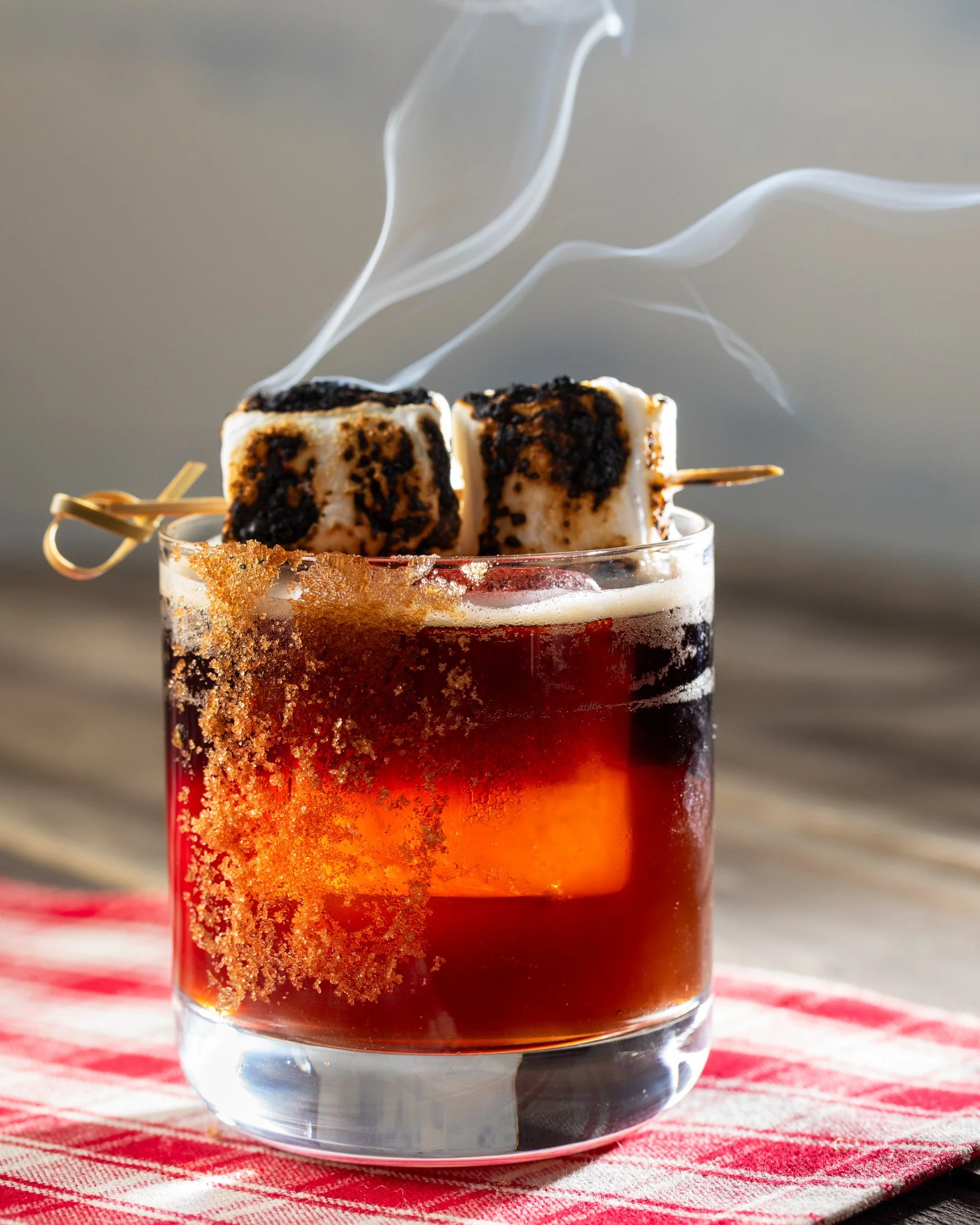 A glass of hot beverage with two toasted marshmallows on skewers, sprinkled with burnt sugar, with steam rising, placed on a red checkered cloth.