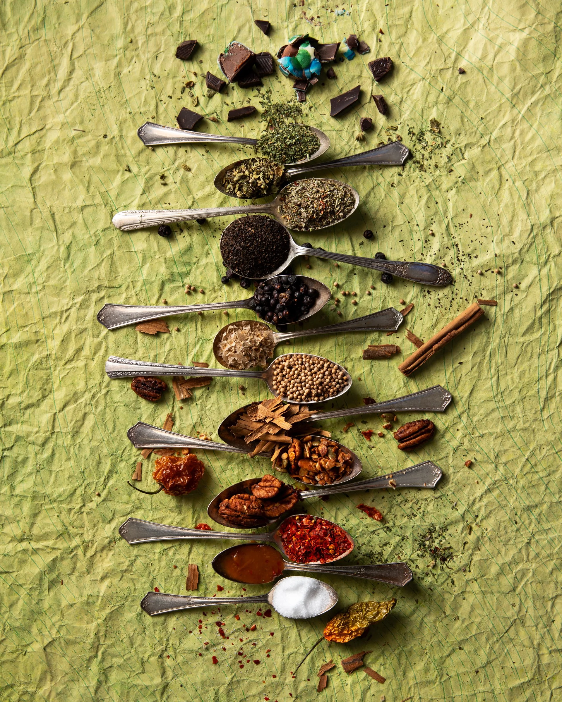 Multiple spoons filled with various spices, herbs, and seasonings arranged on crinkled green paper, with some broken chocolate pieces, candies, and dried herbs scattered around.