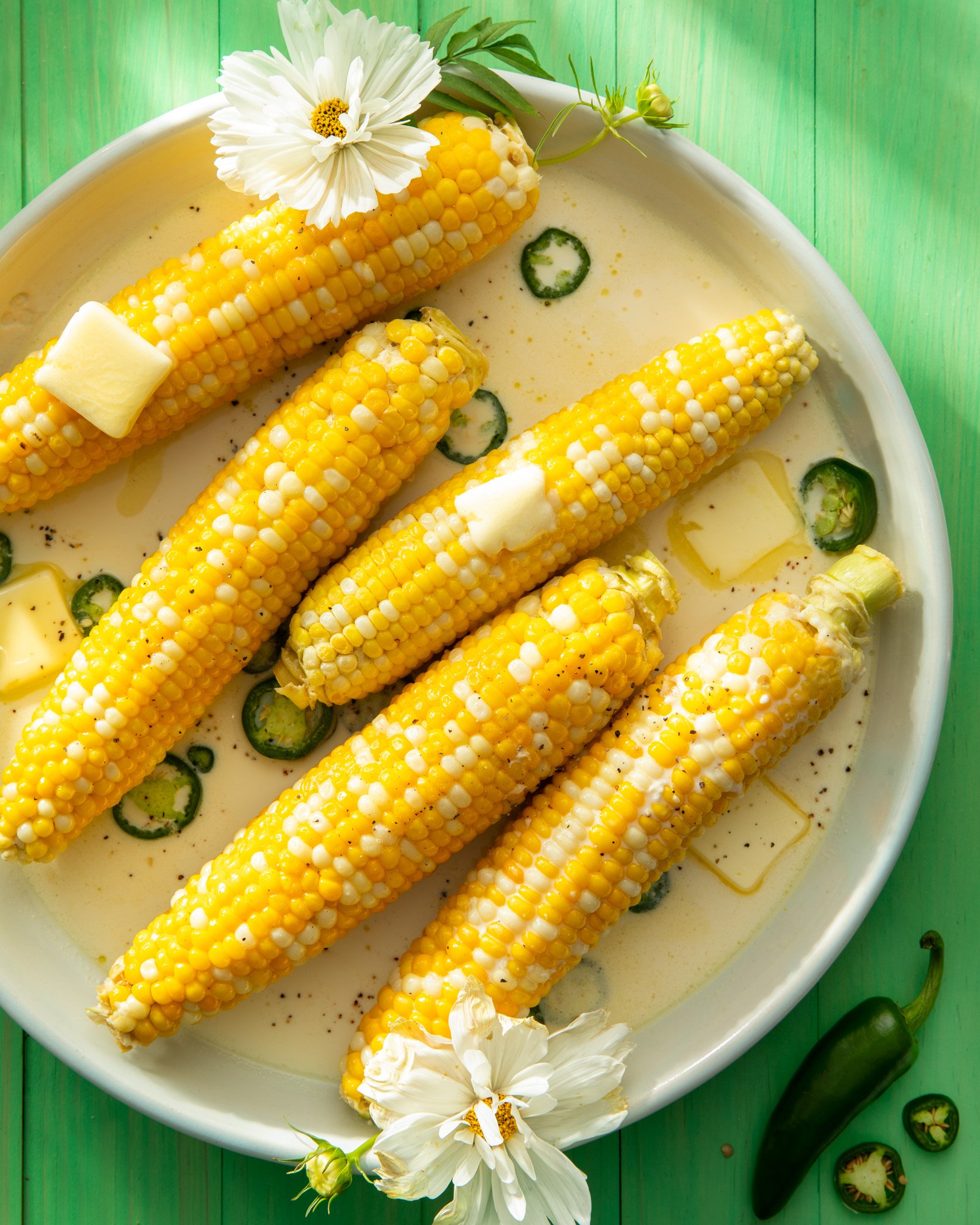 Five ears of corn in a bowl with creamy sauce, slices of jalapeño peppers, and small butter cubes, garnished with white flowers, on a bright green wooden surface.