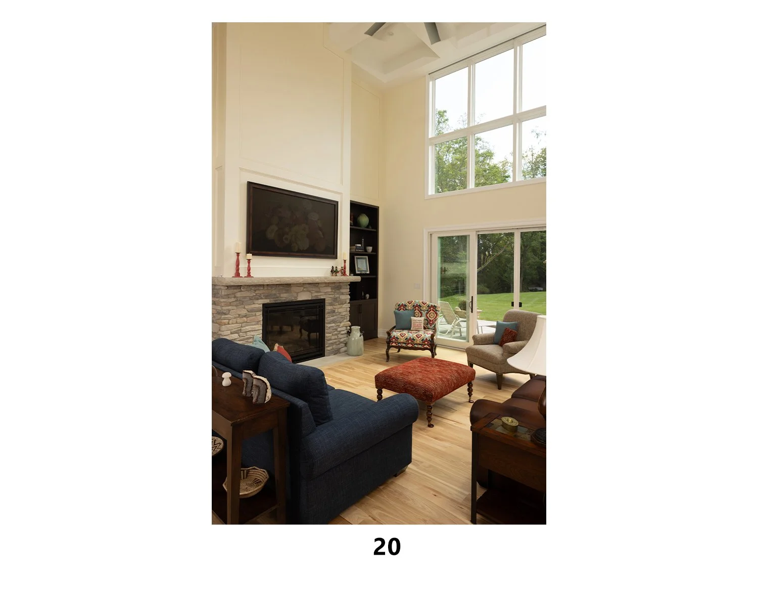 Living room with high vaulted ceiling, large windows, a stone fireplace, a mounted flat-screen TV, and assorted chairs and furniture facing the fireplace.
